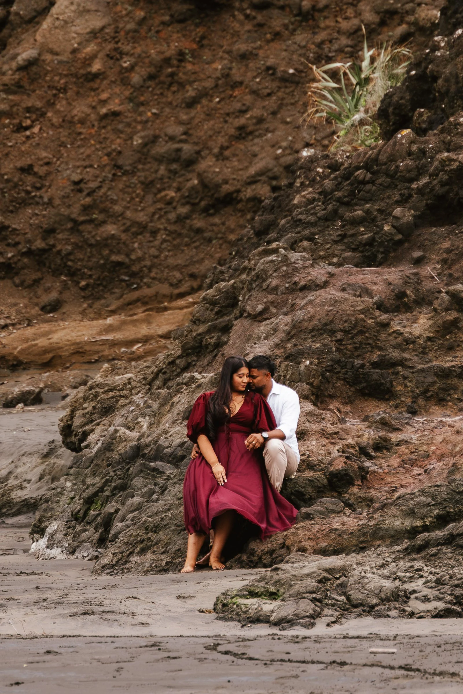 A couple sitting on rocks by the beach, the woman wearing a burgundy dress and the man wearing a white shirt and khaki pants, with a large rock formation behind them.