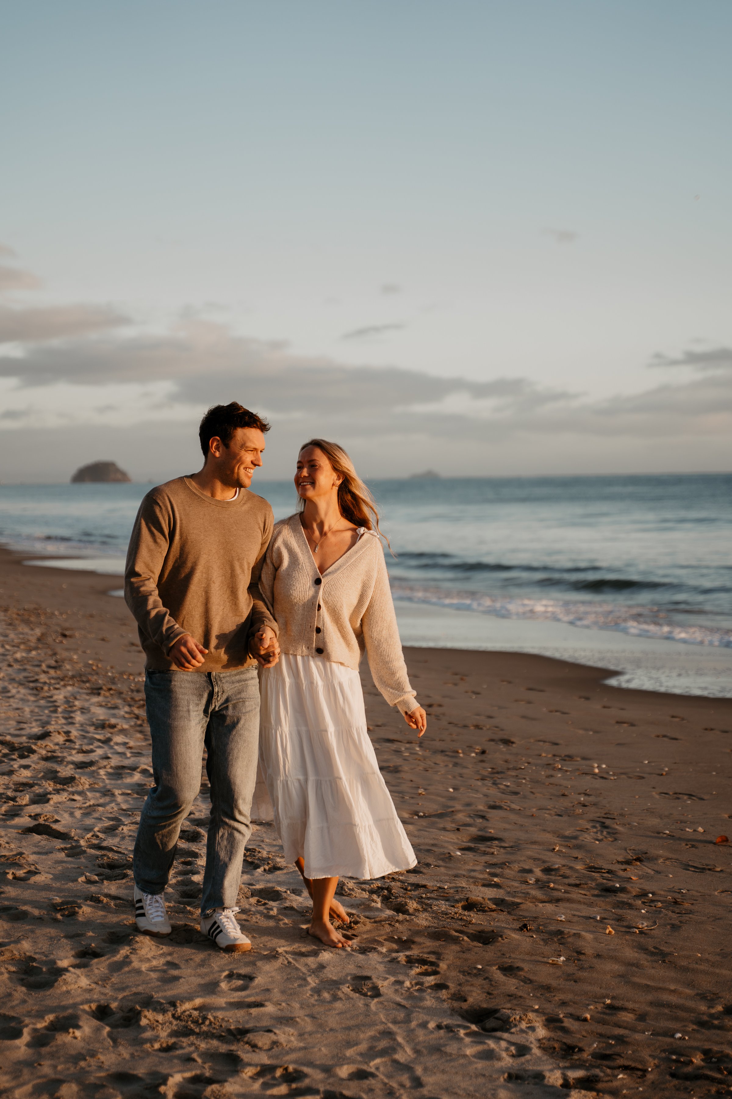 A young couple holding hands and walking along the beach during sunset, with the ocean and sky in the background.