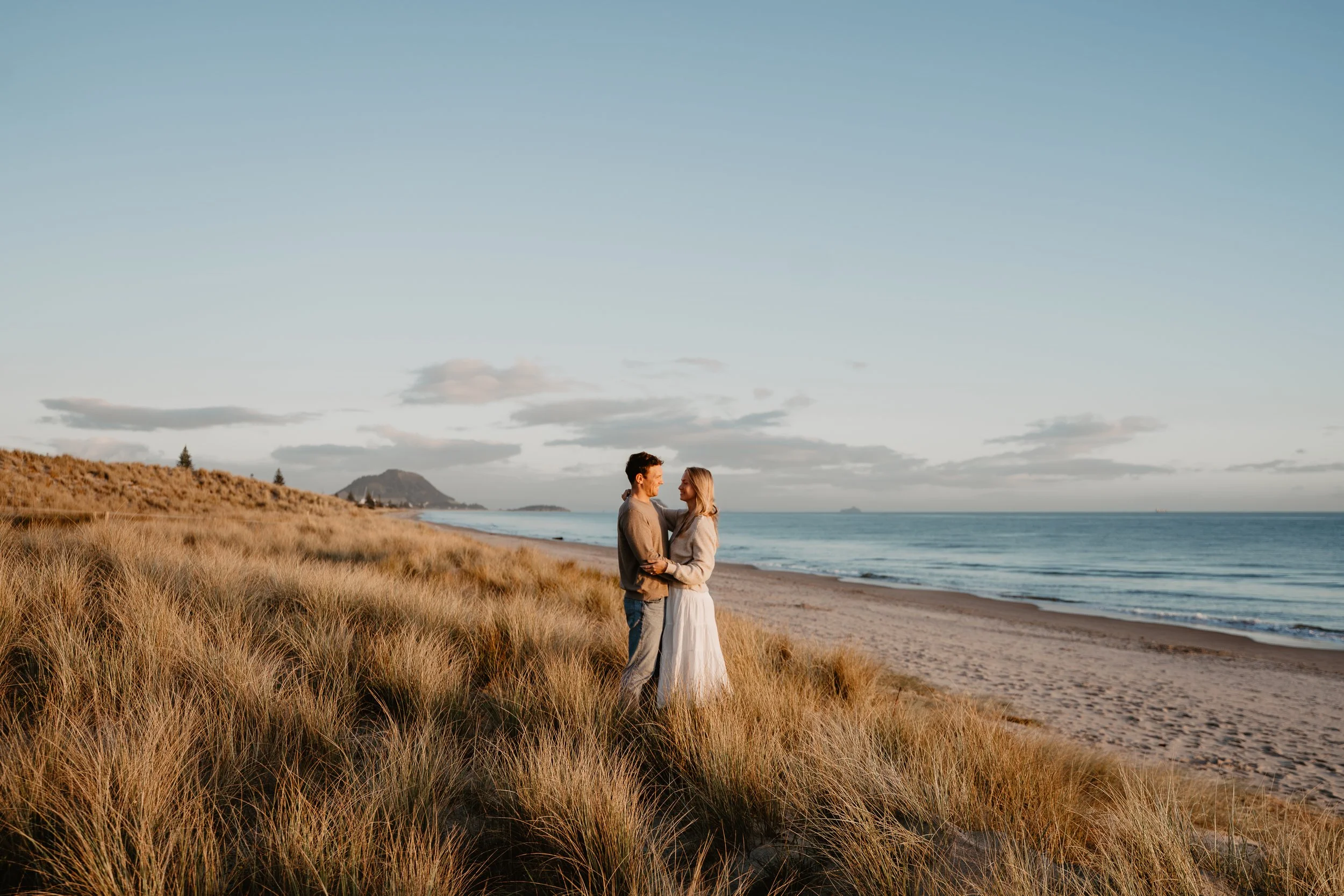 A couple standing on a grassy beach at sunset, facing each other and smiling with arms around each other, near the ocean with cloudy sky and distant landforms in the background.