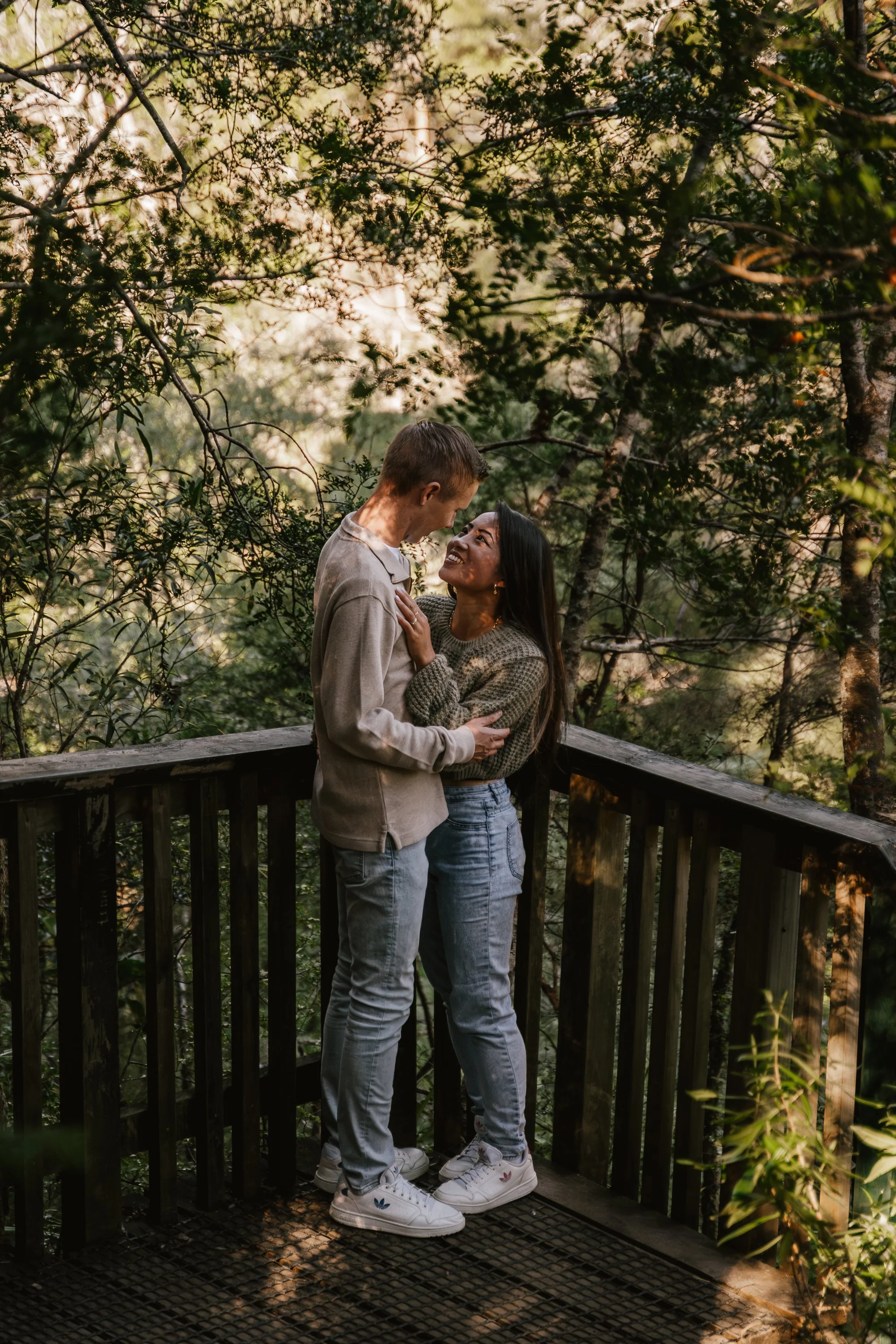 A young couple embracing on a wooden bridge in a forest, smiling and looking into each other's eyes, surrounded by dense green trees and sunlight filtering through the leaves.