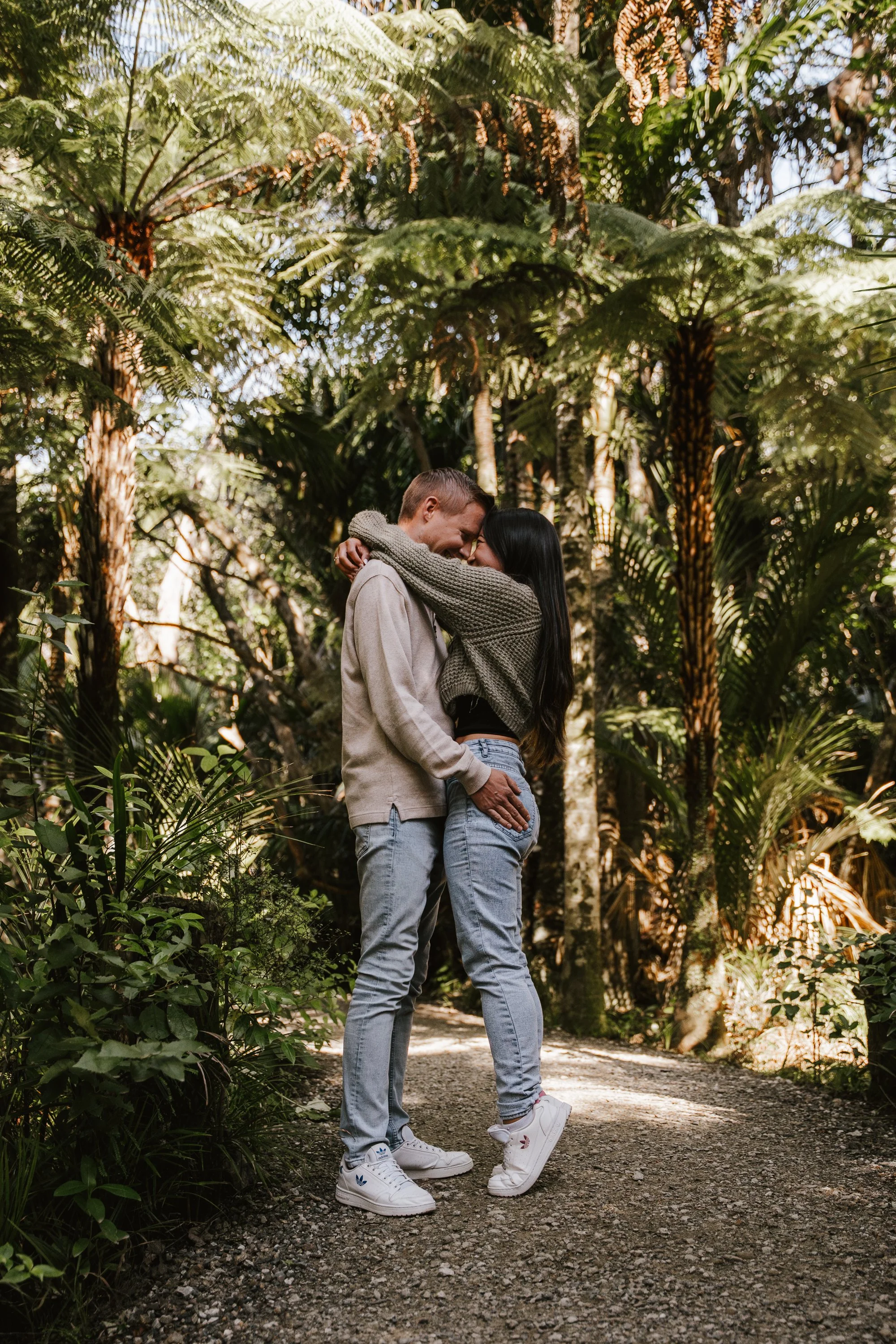 A young couple is embracing and smiling at each other on a dirt path in a lush, green forest with tall trees and dense foliage.