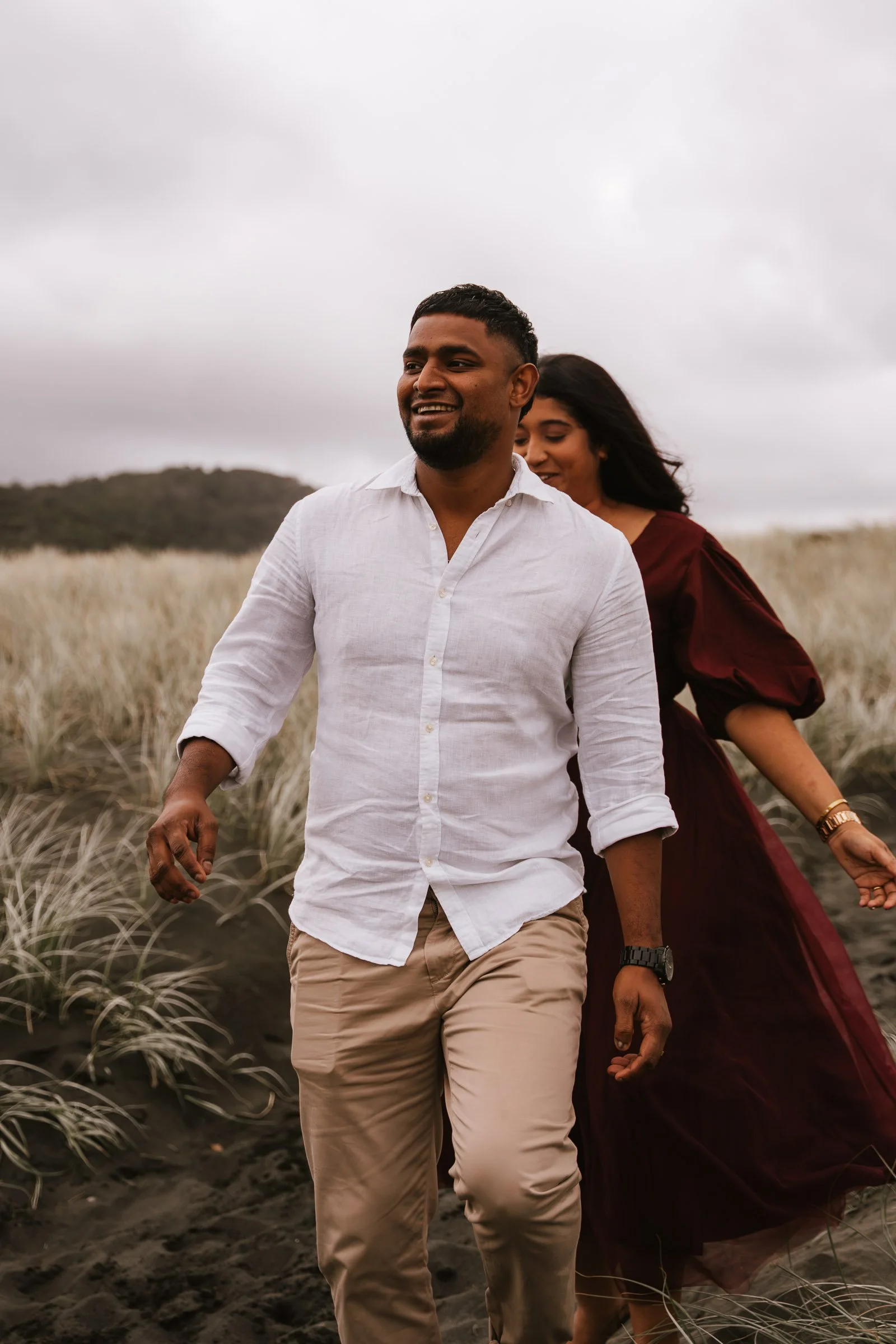 Smiling man in a white shirt and khaki pants walking through tall grass with a woman in a maroon dress behind him, both outdoor setting under cloudy sky.