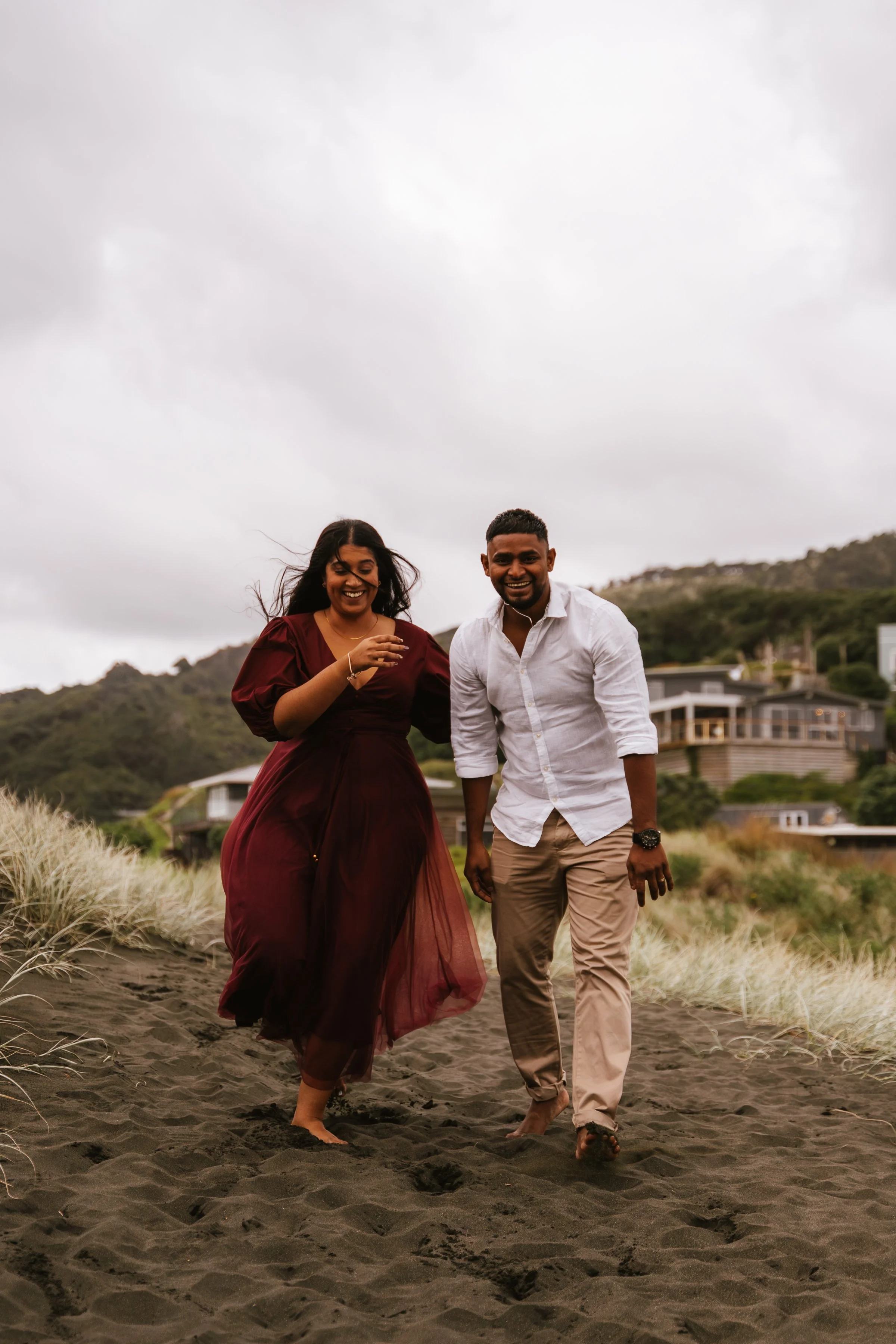 Two smiling friends, a woman in a burgundy dress and a man in a white shirt and beige pants, walk barefoot on a sandy beach with grassy dunes behind them during overcast weather.