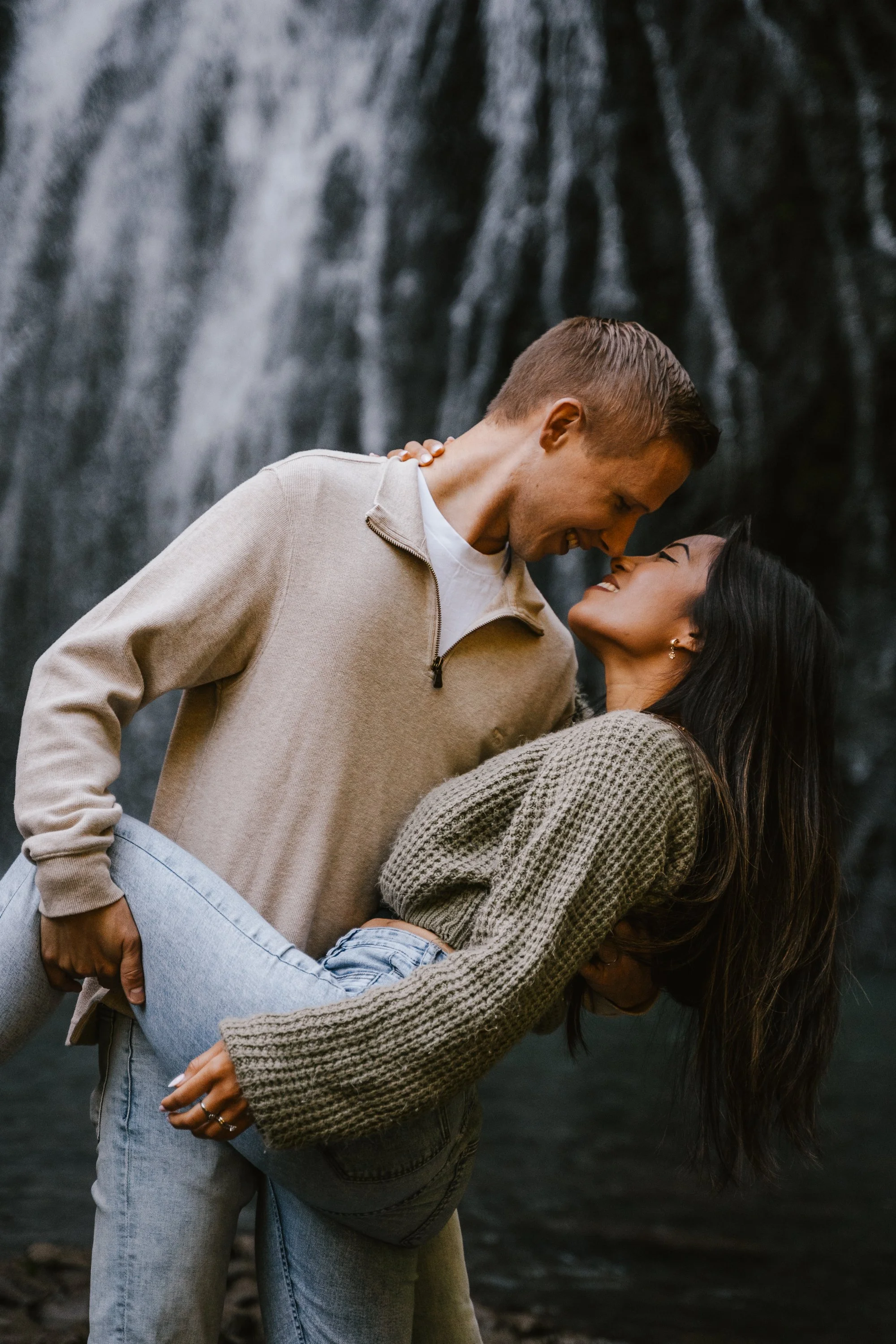 A couple dancing close by a waterfall. The man is smiling and leaning towards the woman, who is looking up and smiling back. They are hugging, and the woman has her arms around the man's neck. The woman is wearing a knitted sweater and jeans, while the man is dressed in a beige sweater and light blue jeans.