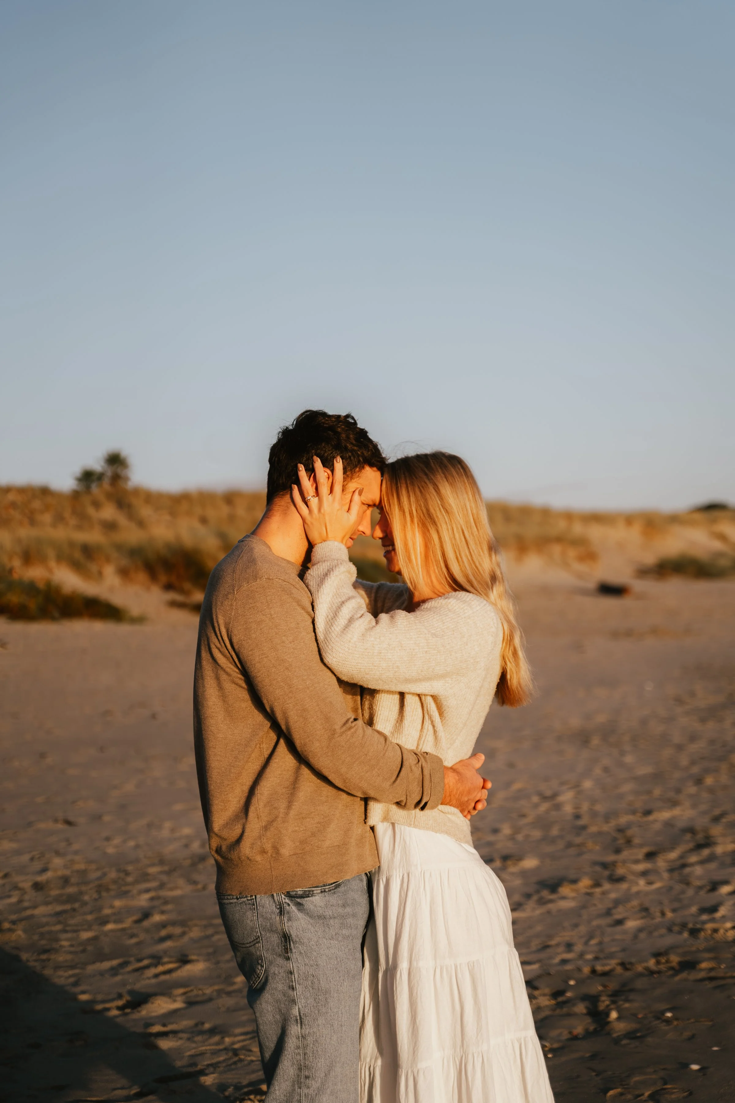A couple, a man and a woman, embracing and touching foreheads on a beach at sunset, with sandy ground and some distant vegetation in the background.