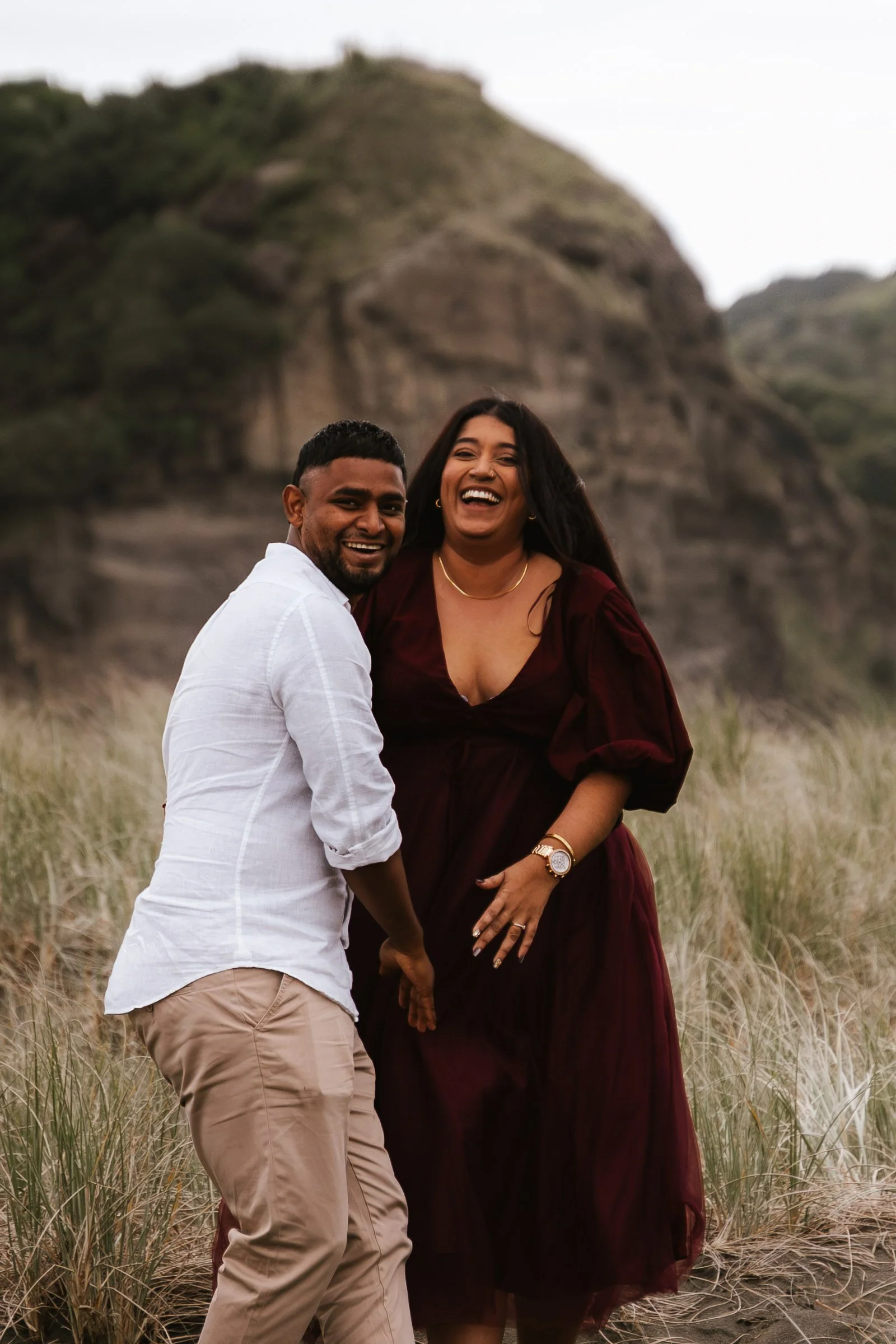 A smiling man and woman standing close together outdoors in a grassy field with rocky hills in the background, facing the camera.