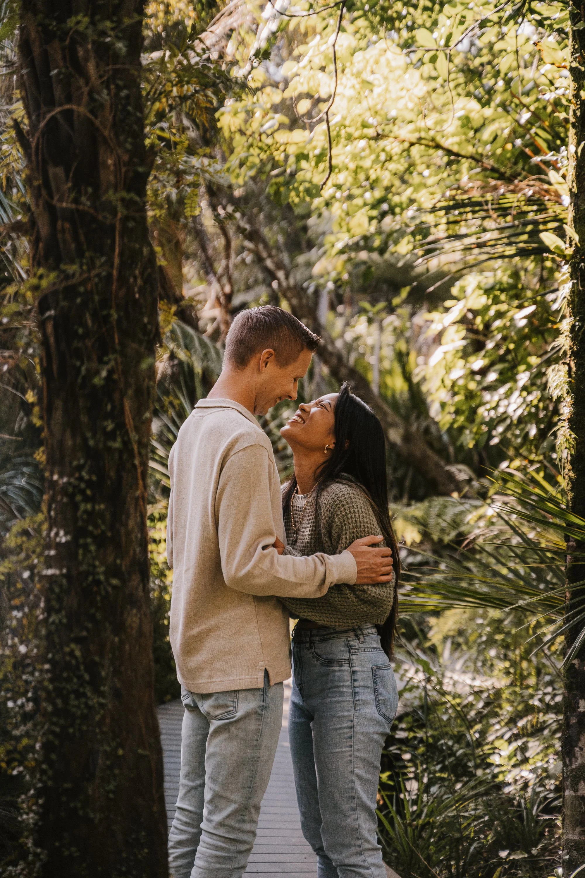 A couple stands close and smiles at each other in a lush, green outdoor setting with trees and foliage.