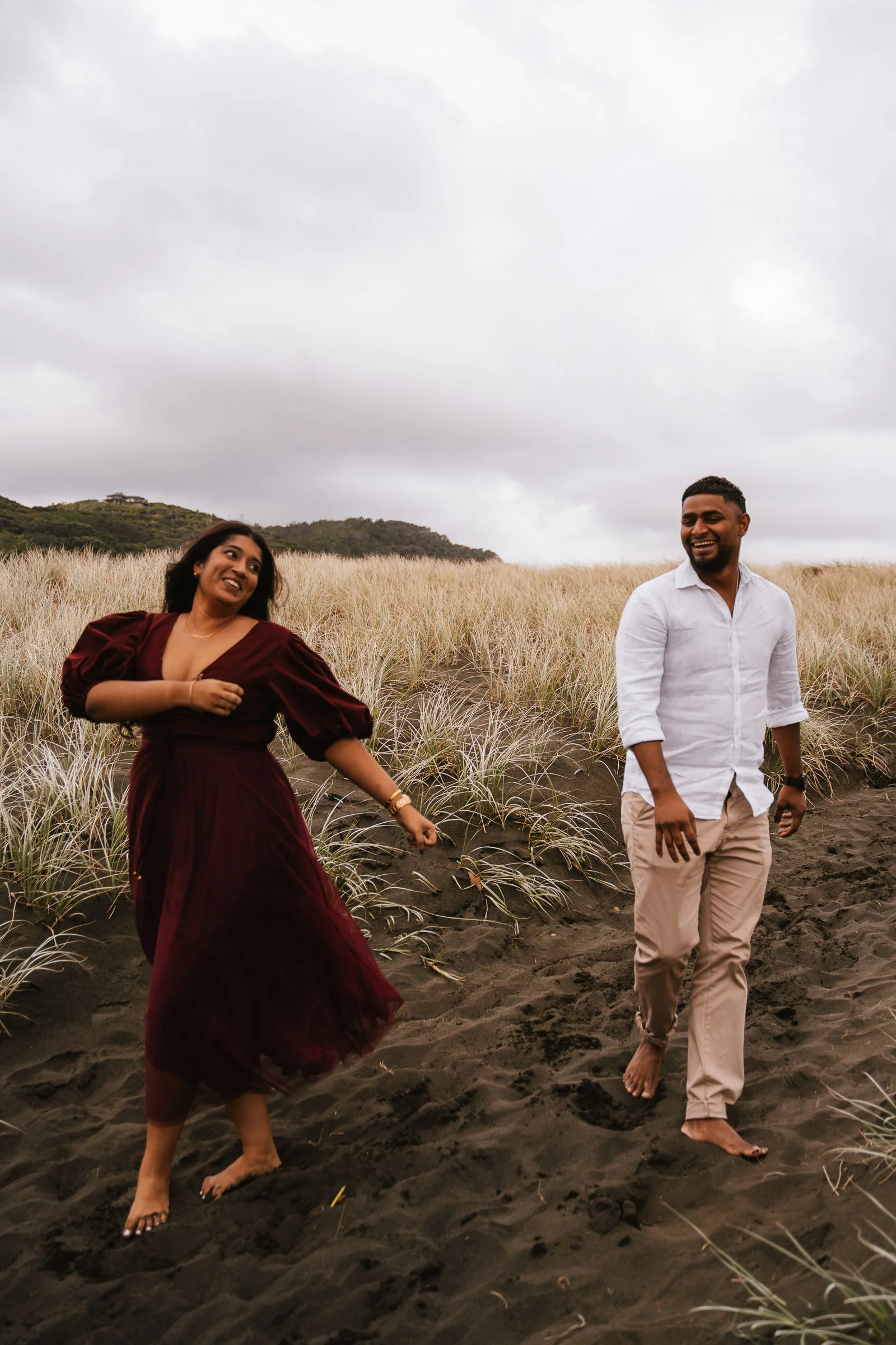 A woman and a man walking barefoot on a sandy beach; the woman is wearing a dark red dress and the man is dressed in a white shirt and beige pants; they are smiling and looking happy, with high grass and hills in the background under a cloudy sky.