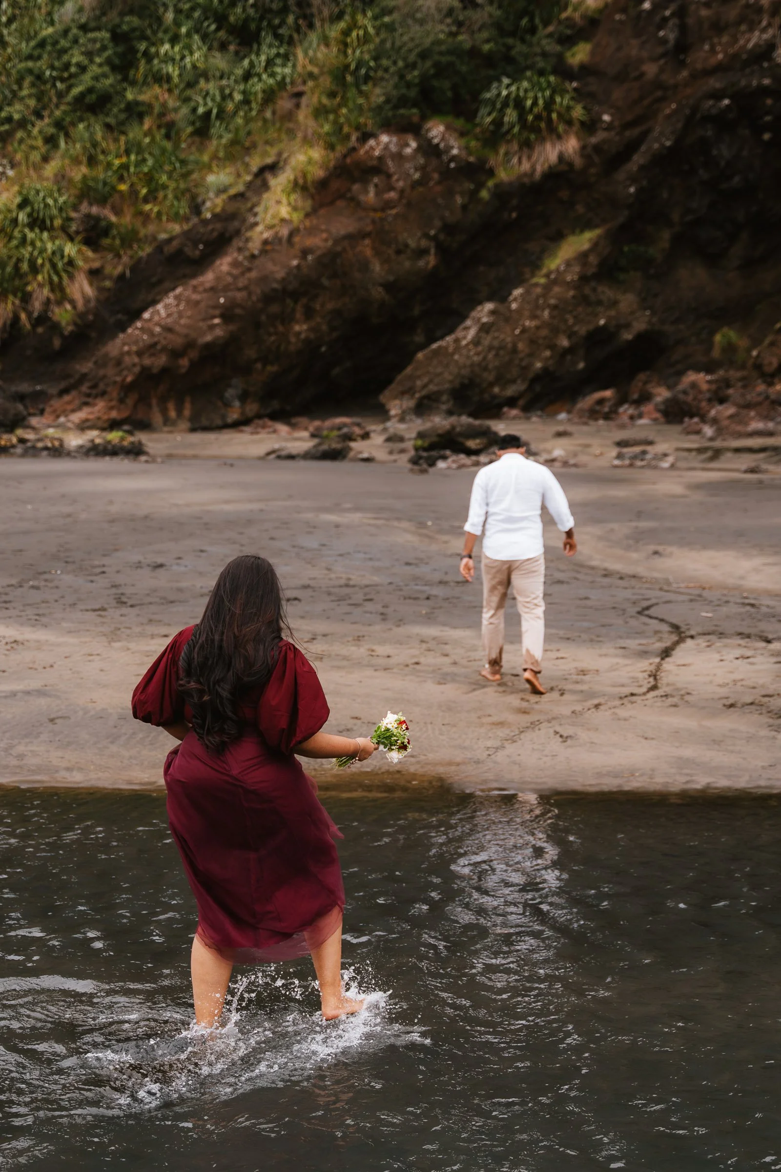 A woman in a burgundy dress stands in water, holding a bouquet, while a man in beige pants and a white shirt walks forward on a sandy beach near rocky cliffs and greenery.