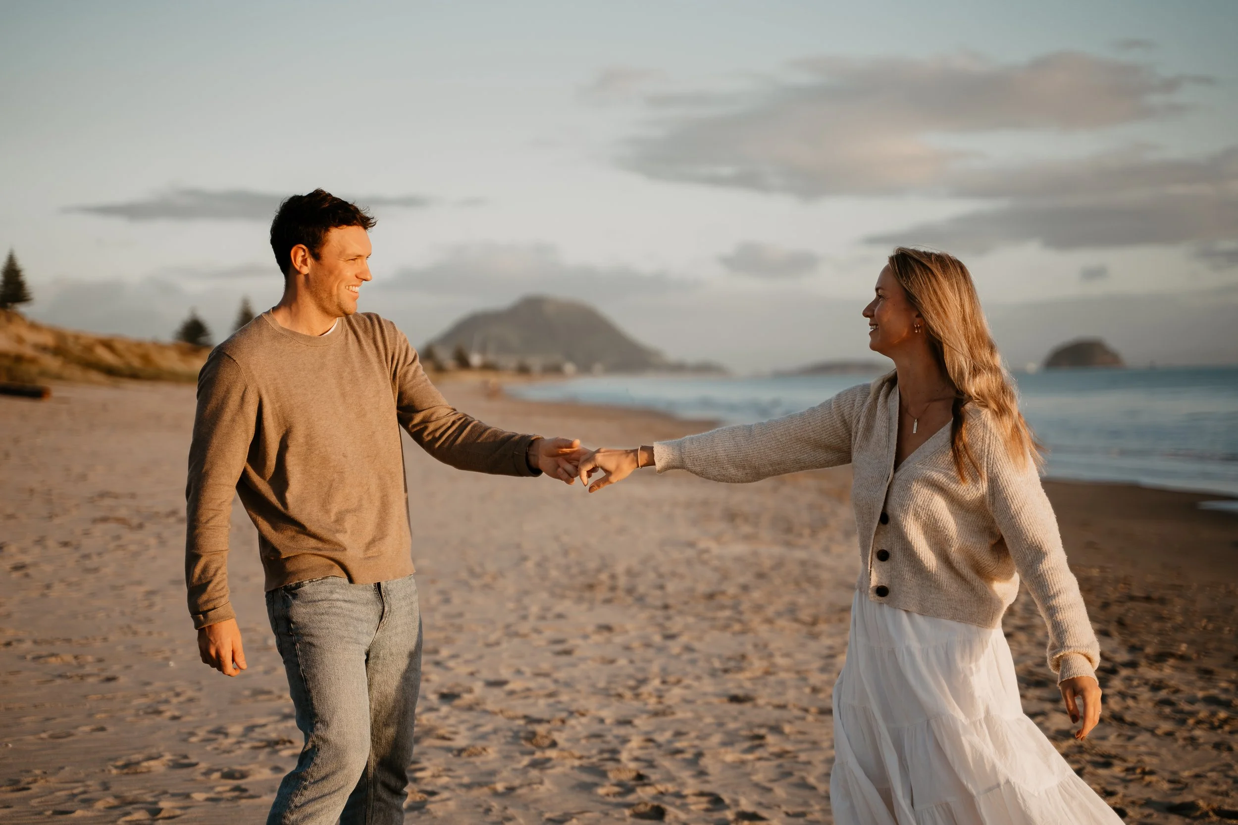 A smiling man and woman holding hands on a beach during sunset, with a mountain and ocean in the background.