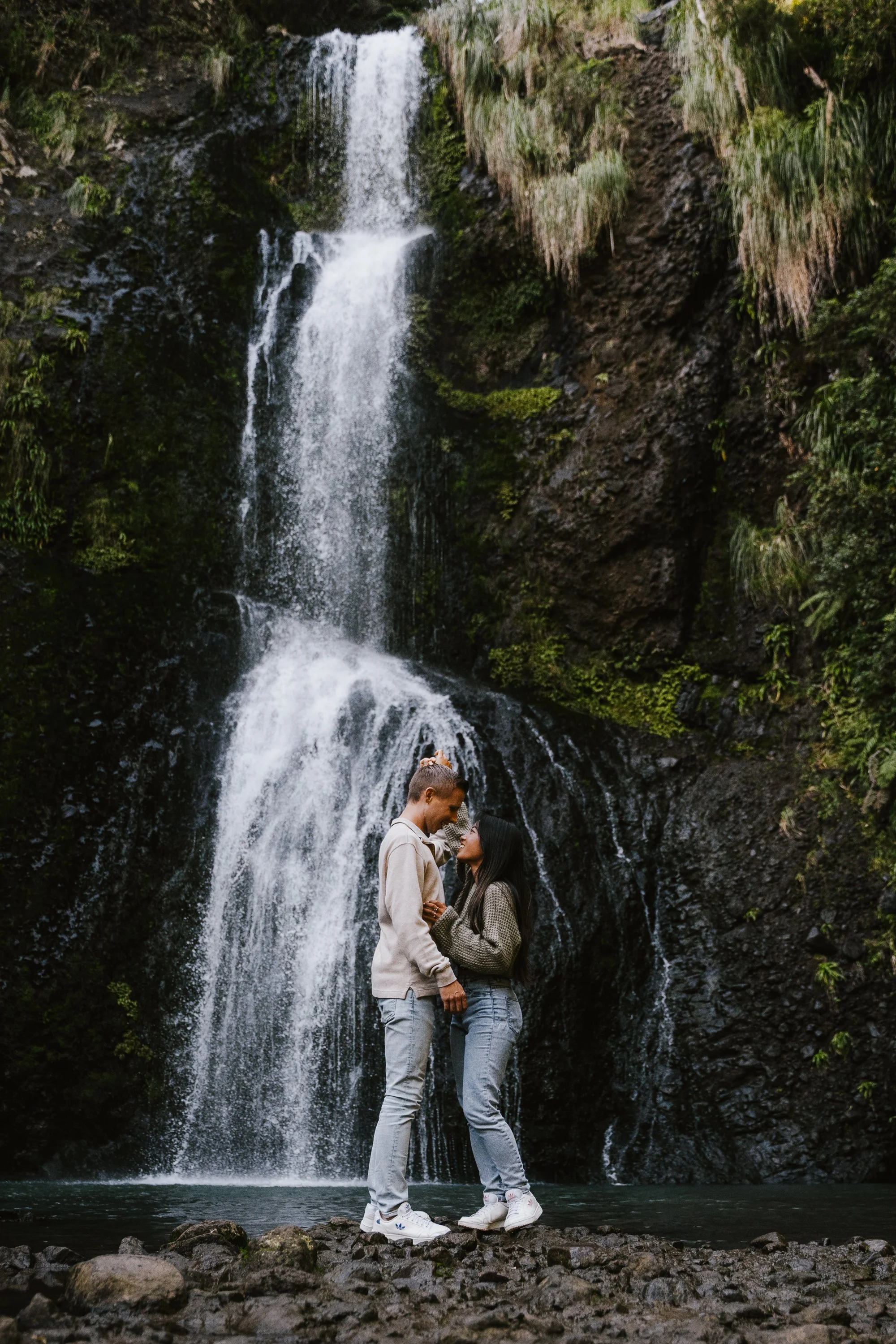A couple stands in front of a waterfall, holding hands and gazing at each other, with lush greenery surrounding the waterfall.