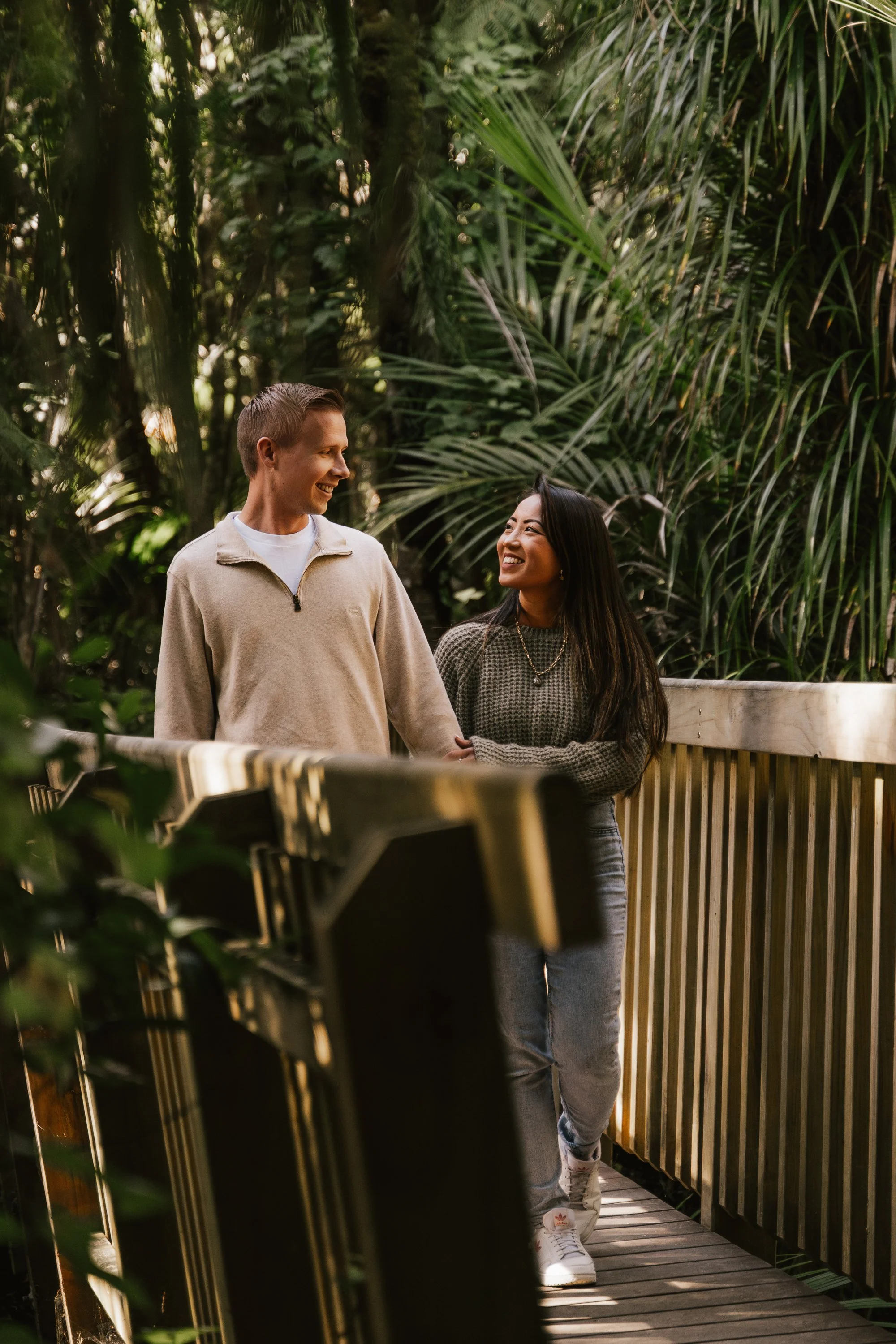 A young man and woman holding hands and smiling at each other on a wooden walkway surrounded by lush green foliage.