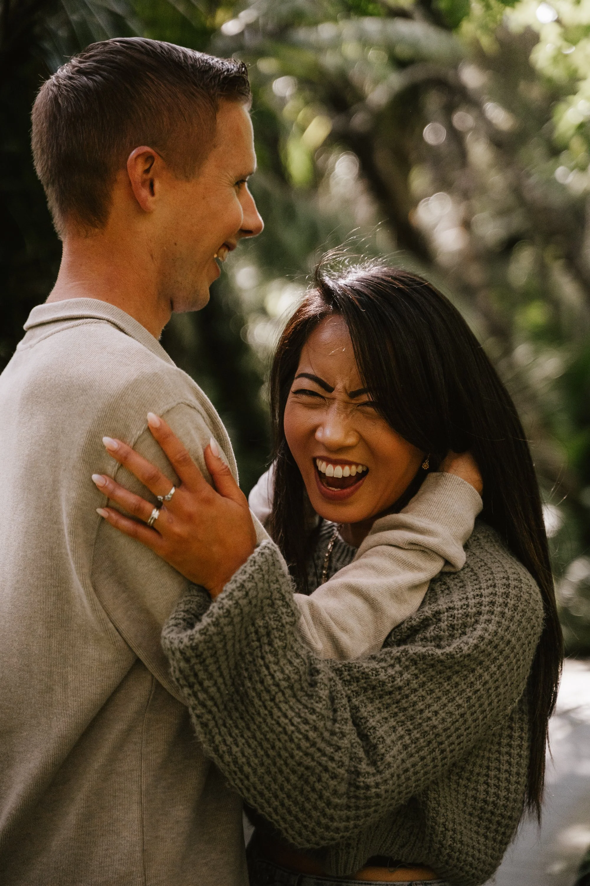 A man and woman are smiling and embracing outdoors among trees, with a relaxed and joyful interaction.