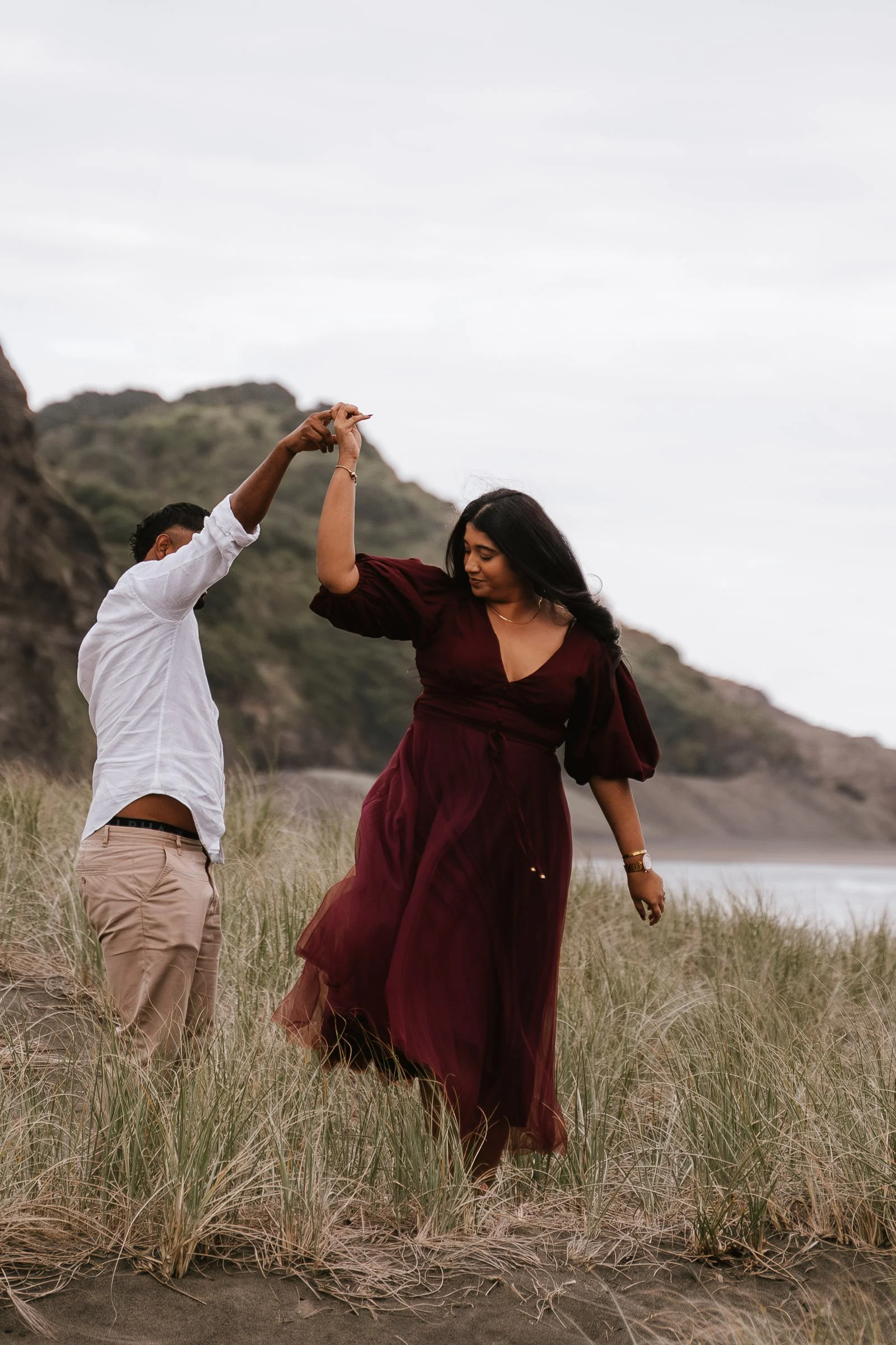 A couple dancing on a sandy beach surrounded by tall grass, with mountains and cloudy sky in the background.