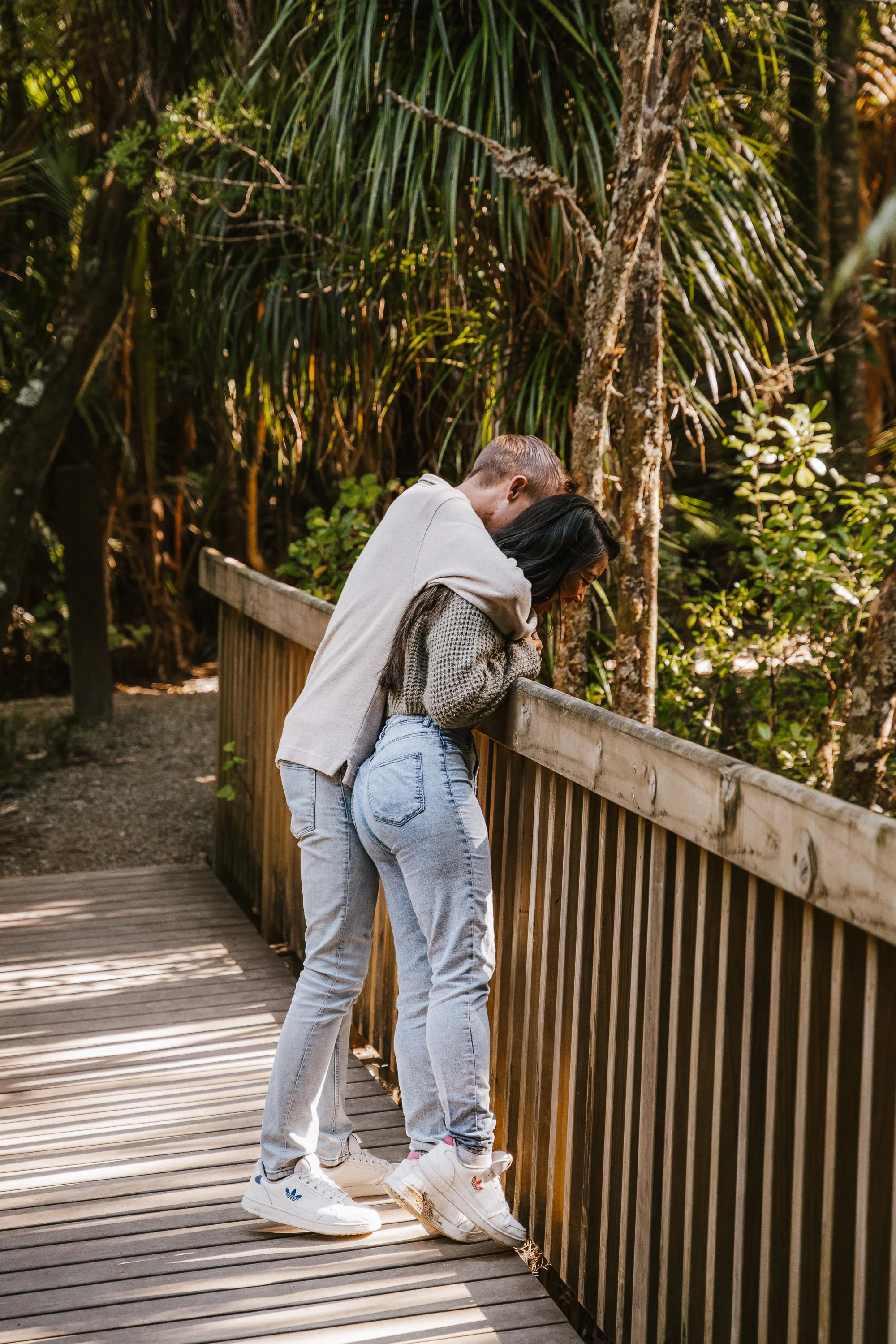 Two people standing close together on a wooden bridge in a forest, looking down at something and sharing a moment of closeness.