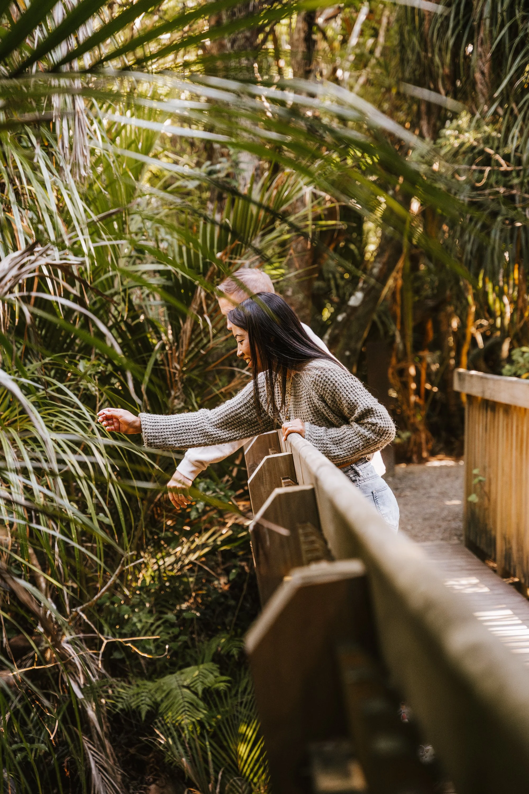 Two women are leaning over a wooden railing, surrounded by dense green jungle foliage, as they observe or interact with the plants.