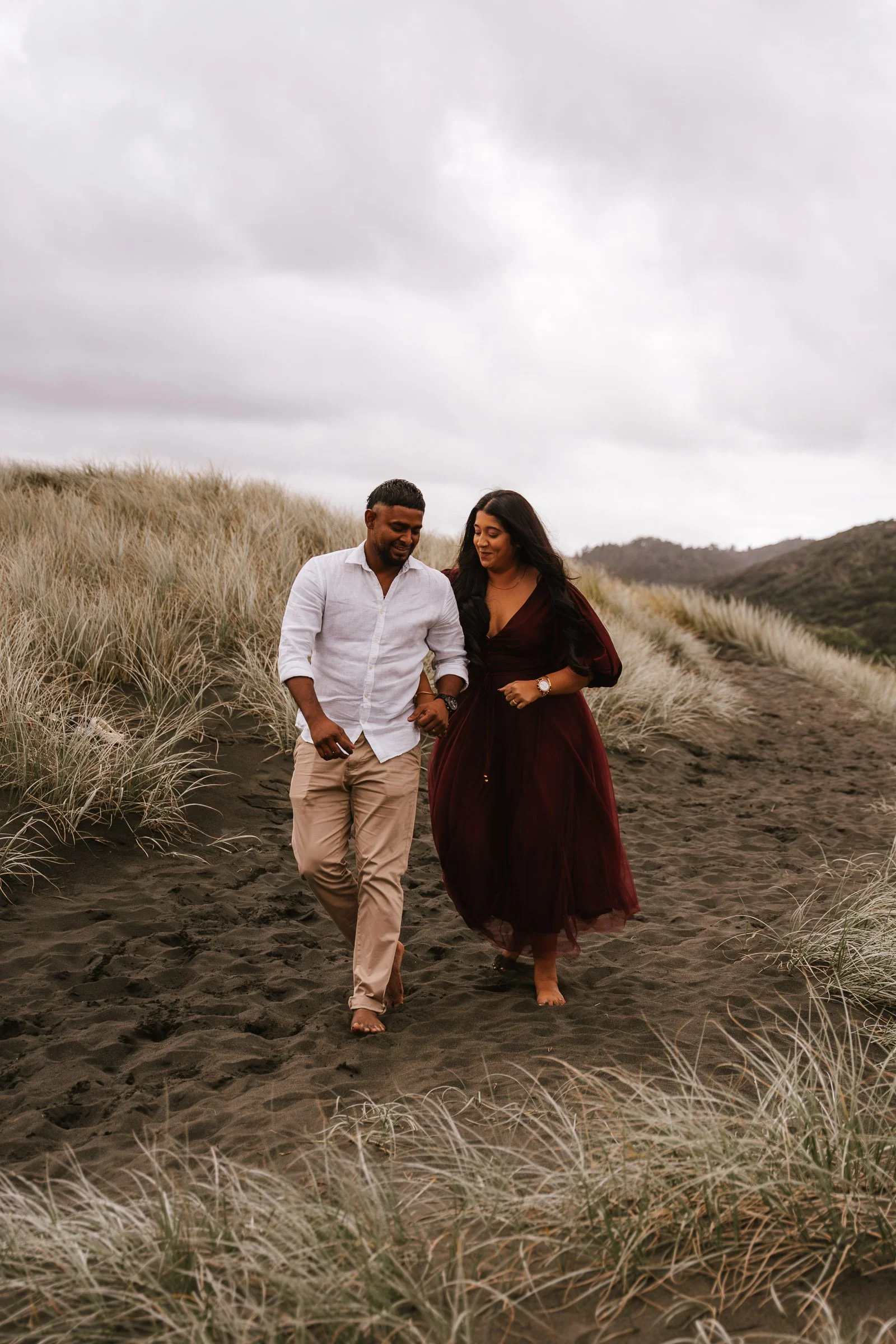A couple walking barefoot on a sandy trail surrounded by tall grass, with overcast sky and hills in the background.