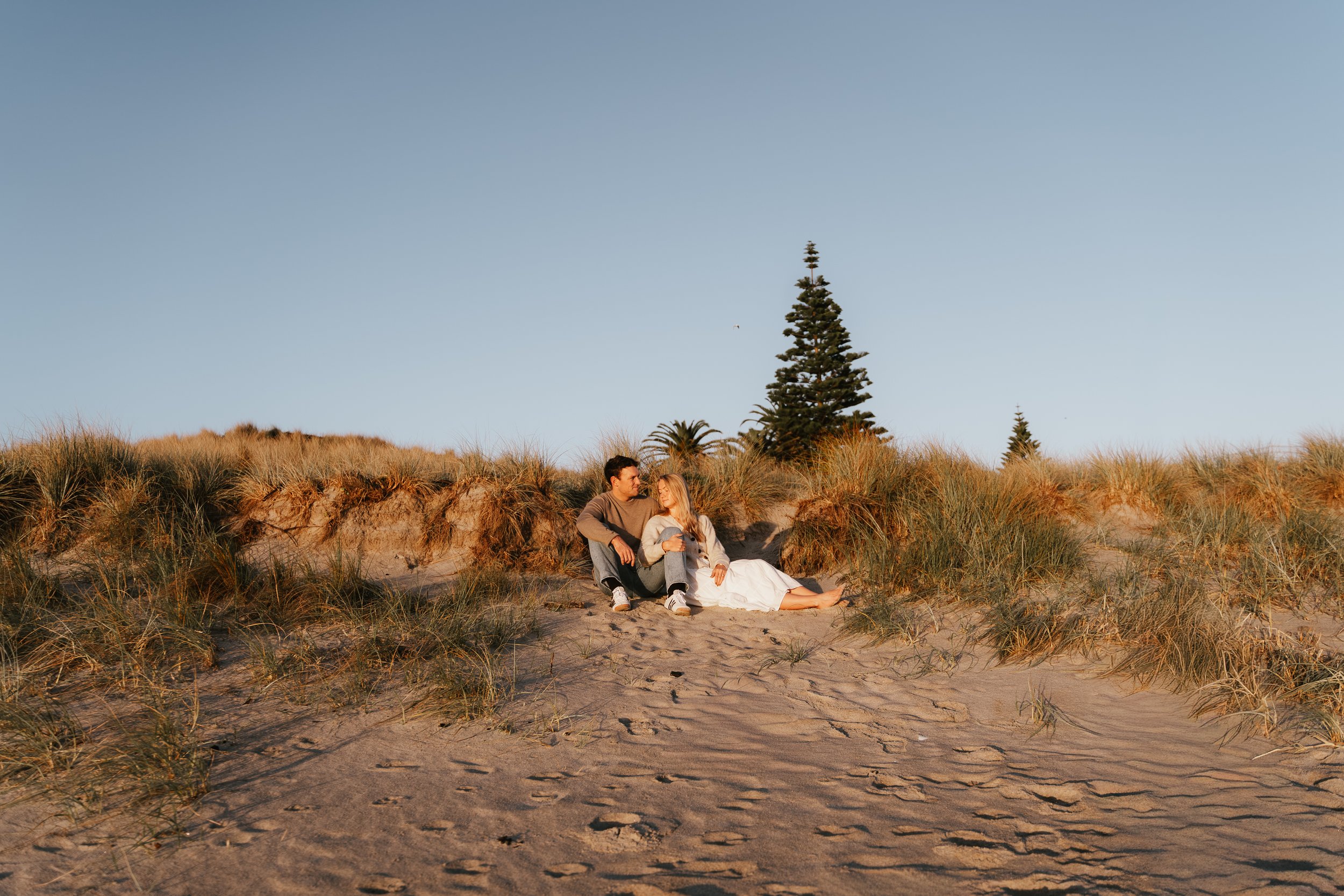 A couple sitting on the sandy beach among grassy dunes during sunset, with tall trees and a clear sky in the background.