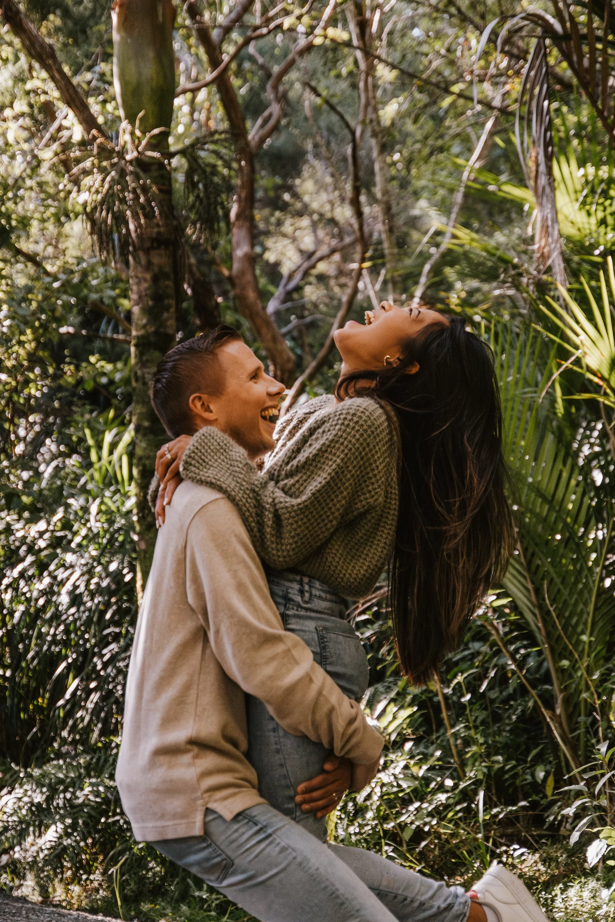 Two young people, a man and a woman, are enjoying a joyful moment outdoors among lush green tropical plants. The woman is being lifted by the man, both smiling and laughing. The woman has long dark hair, and the man has short brown hair.
