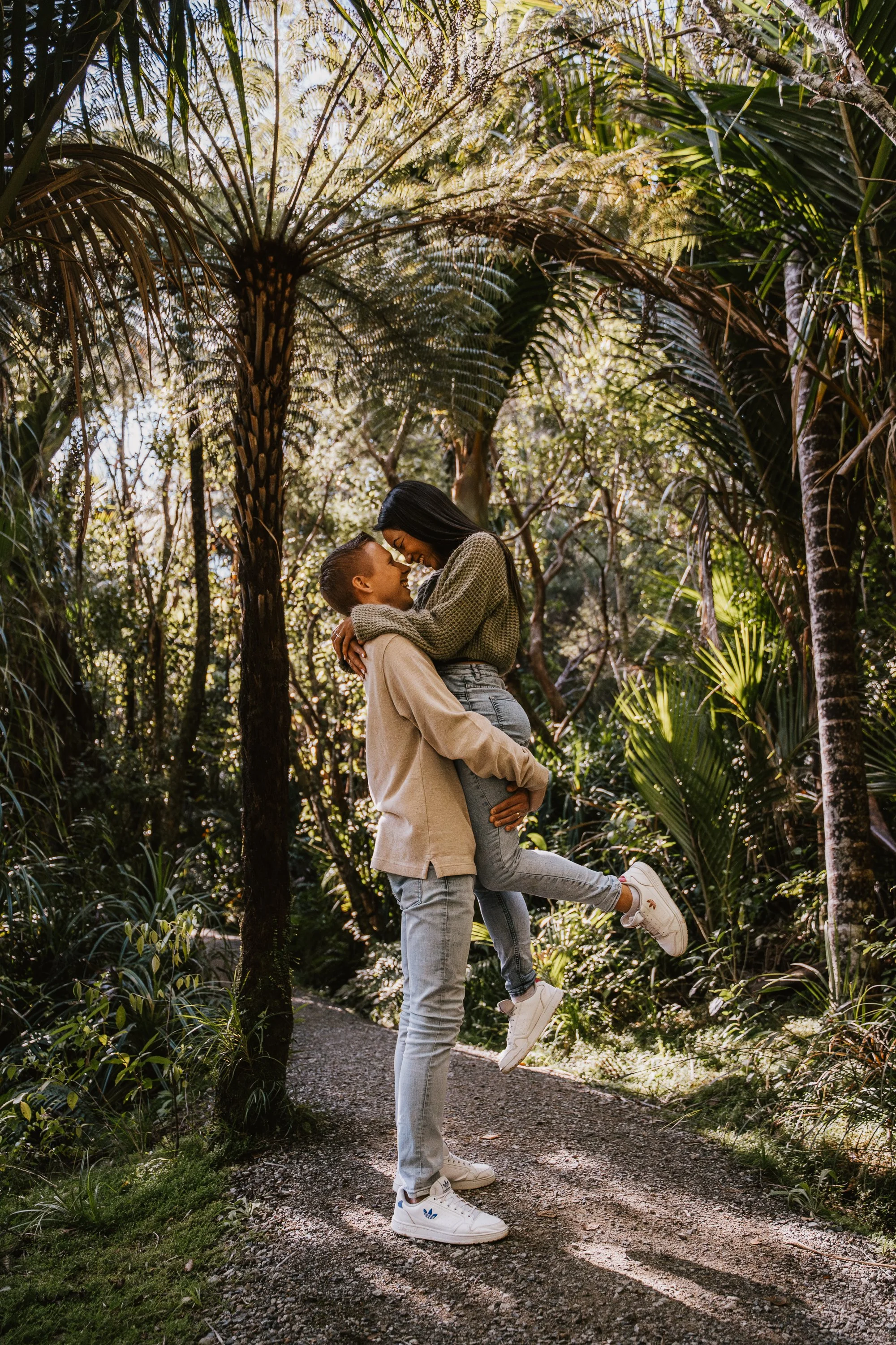 A young couple enjoying an outdoor moment in a lush forest, with the man lifting the woman and both smiling at each other, surrounded by tall tropical trees and greenery.