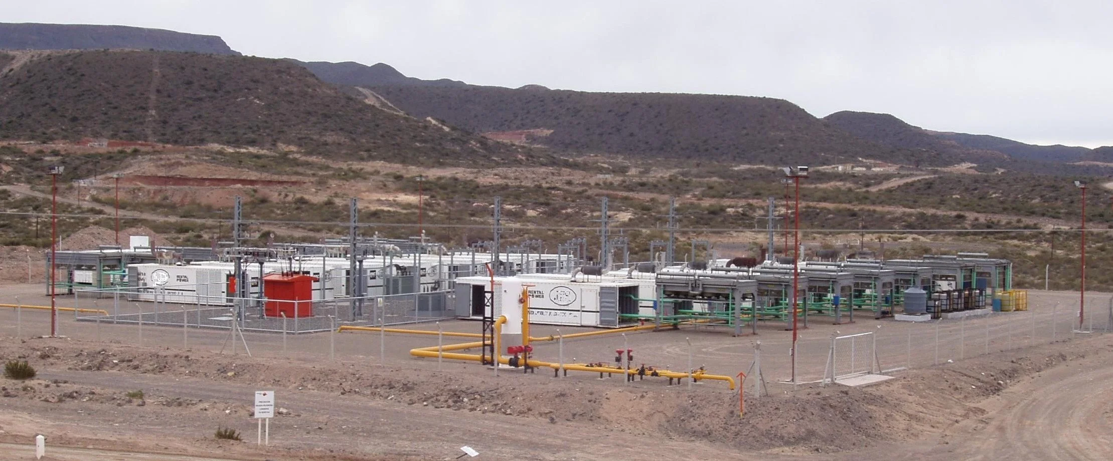 An industrial facility with metal structures, pipelines, and equipment, fenced off in a desert landscape with hills and mountains in the background.