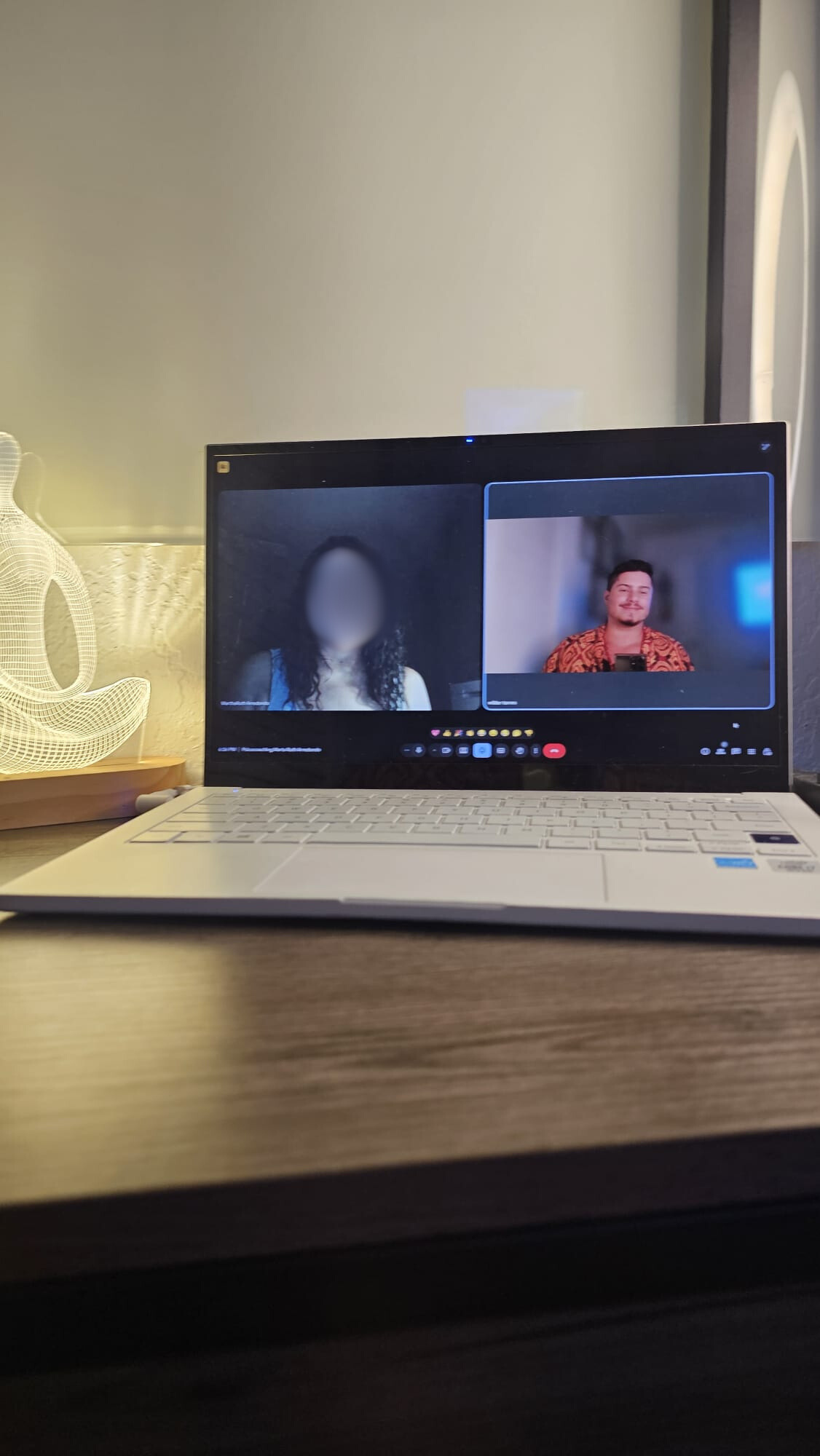 A mobile phone screen showing a video call with the contact name 'Psicocoachingchir'. The screen displays two people in the video call: the main person with a blurred face and a small inset showing another person wearing glasses, smiling. The background in the main video frame features white walls and colorful framed pictures.