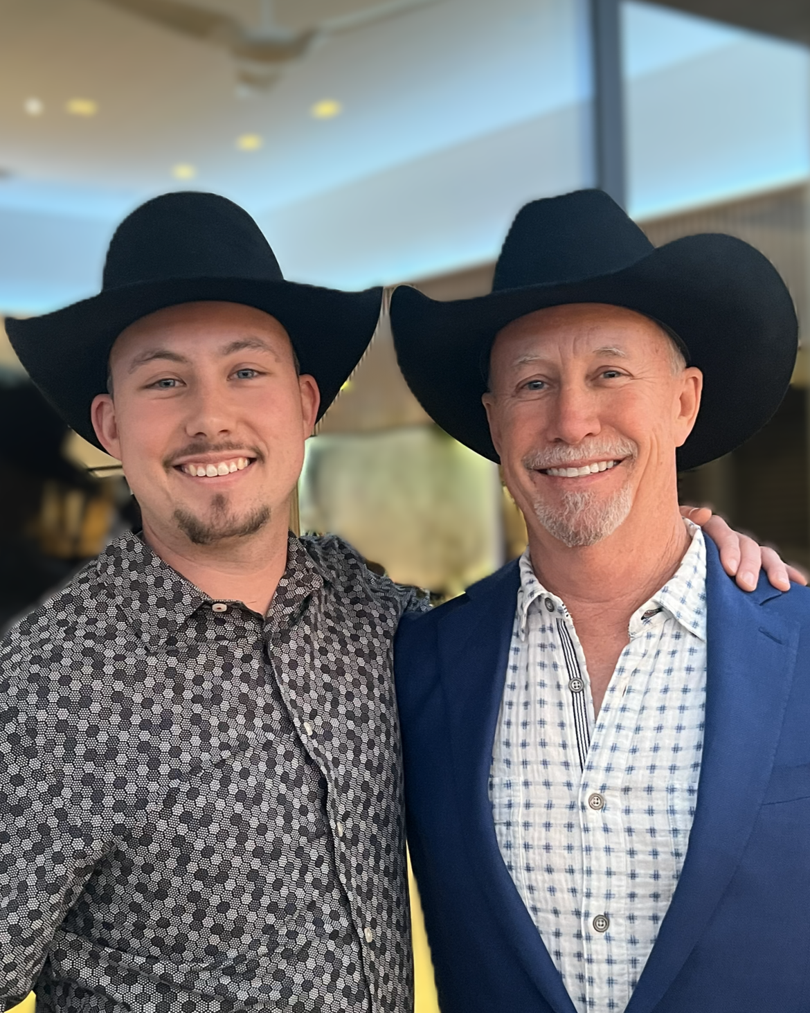 Two men wearing large black cowboy hats, smiling, standing close together indoors. The man on the left has short hair, light facial hair, and wears a patterned button-up shirt. The man on the right has a goatee, short hair, and is dressed in a blue blazer over a checkered shirt. Both are smiling and one has his arm around the other's shoulder.