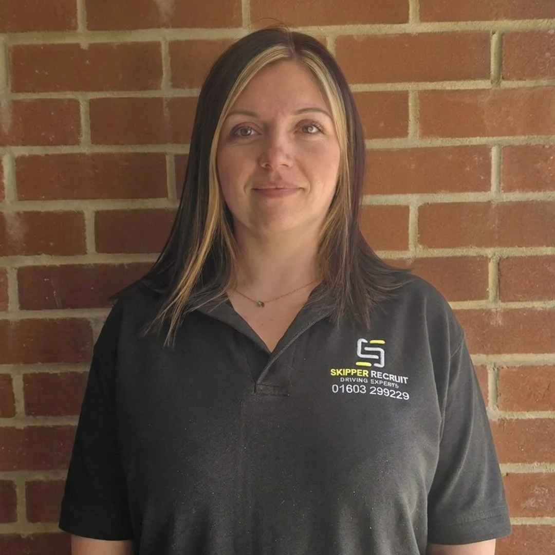 A woman with dark and blonde hair standing against a brick wall, wearing a black polo shirt with a logo and text on the left side.