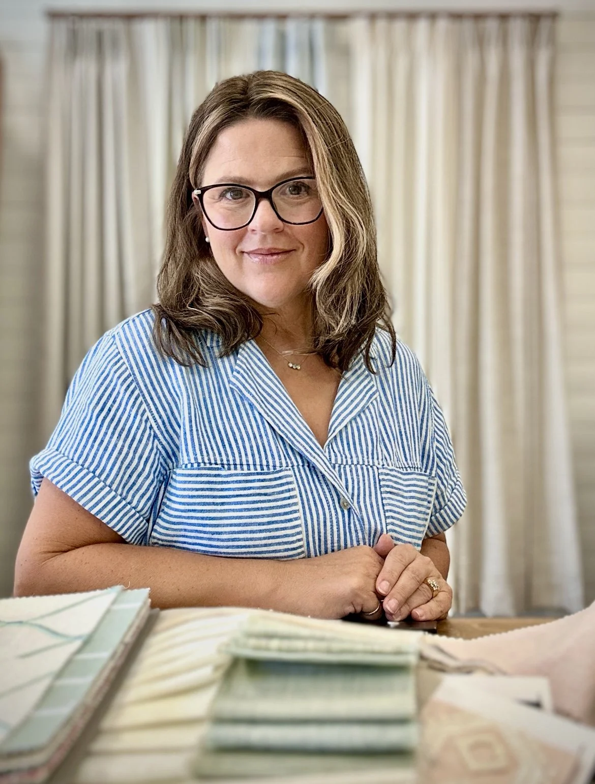 The female owner with glasses and brown hair wearing a blue and white striped shirt sitting at a table with fabric samples, in front of beige curtains.