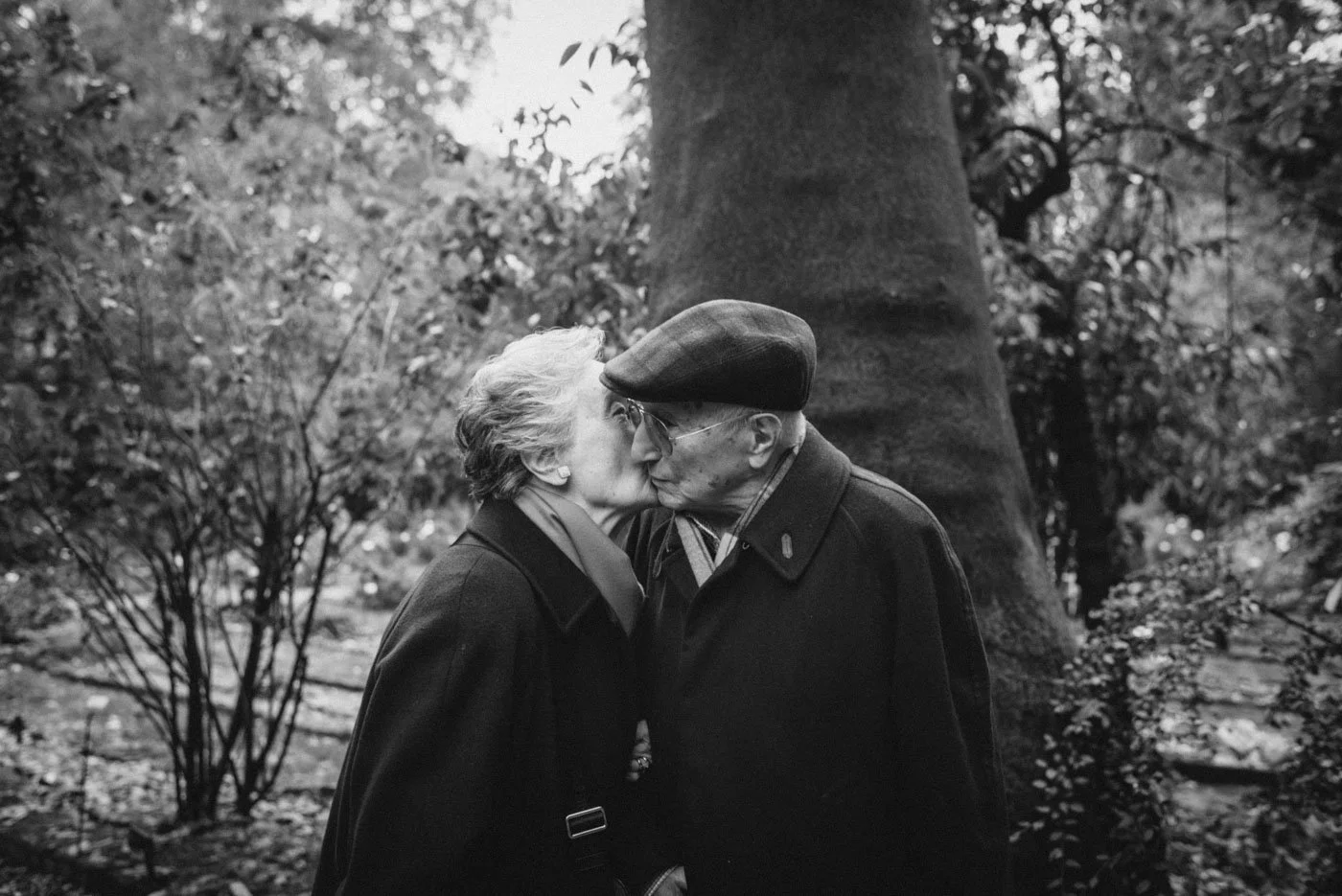 An elderly couple sharing a kiss outdoors in a wooded area, black and white photograph.