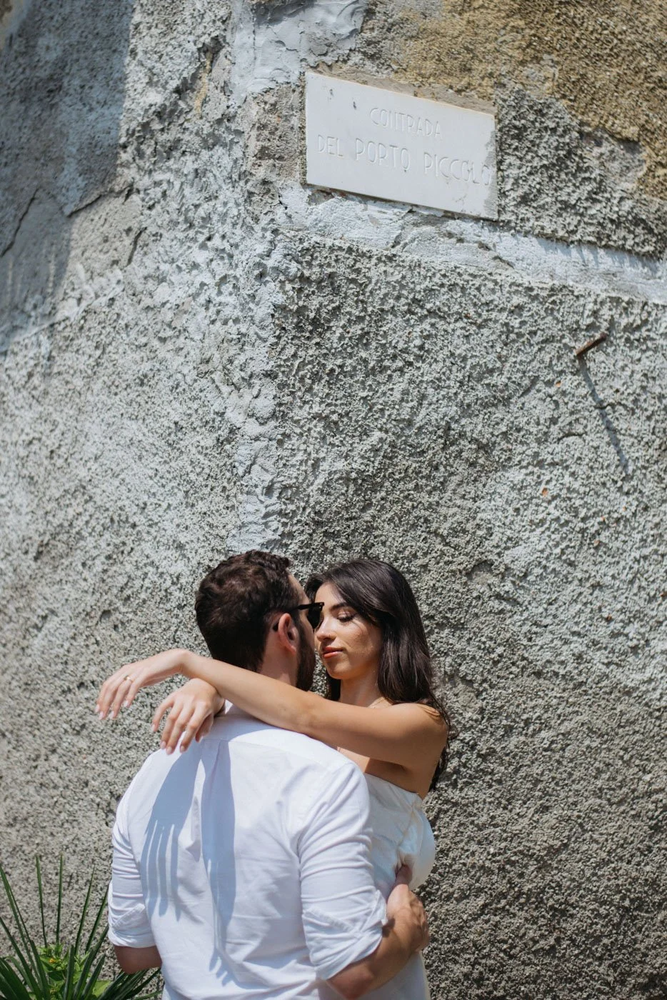 A man and woman embracing in front of a textured stone wall with a sign reading 'CITADA DEL PORTO PICCOLO' in Italian.