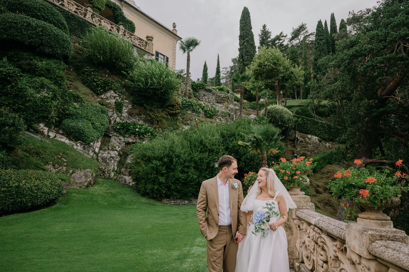 A bride and groom holding hands and smiling at each other on their wedding day in a lush, green garden with various plants and flowers.