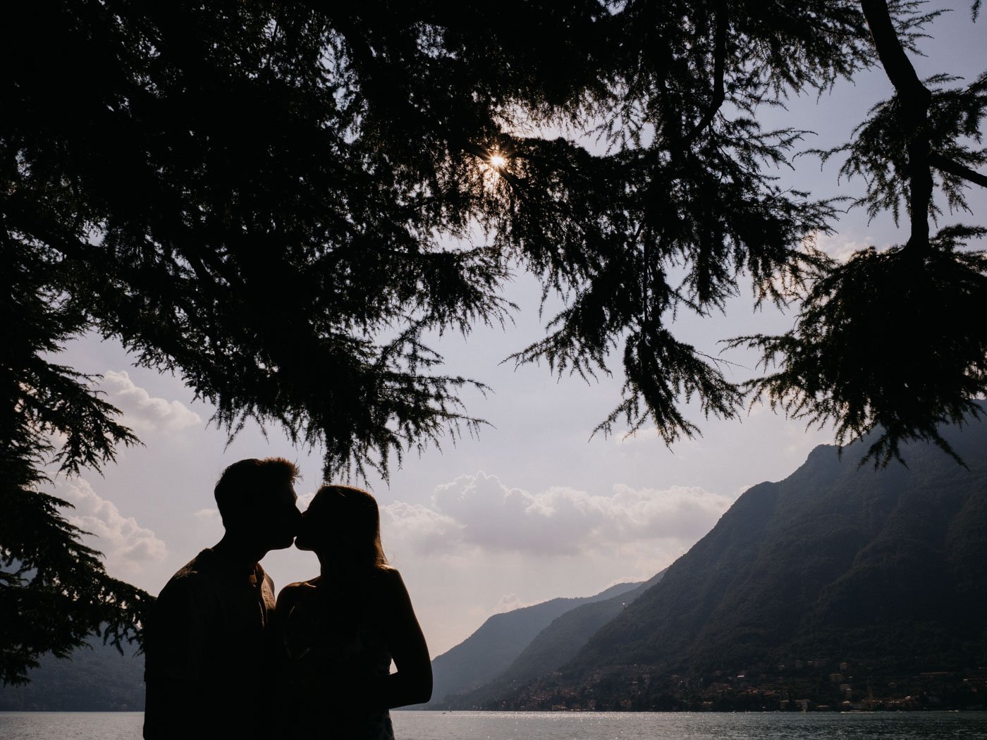 Silhouette of a couple kissing under a tree with mountains, lake, and sky in the background.