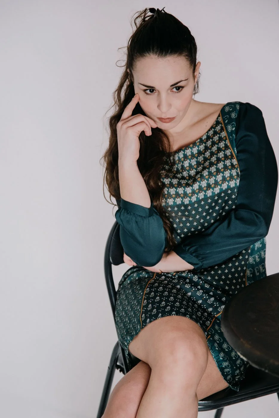 A woman sitting on a chair in a thoughtful pose, resting her chin on her hand, wearing a patterned dress with long sleeves.