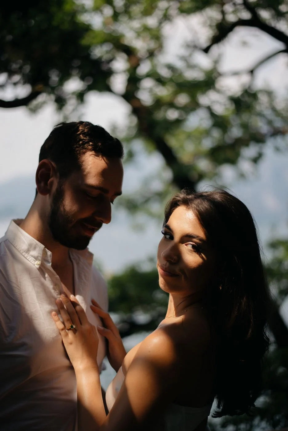 A man and woman standing closely outdoors under a tree, with sunlight casting shadows on their faces, creating a warm and intimate atmosphere.