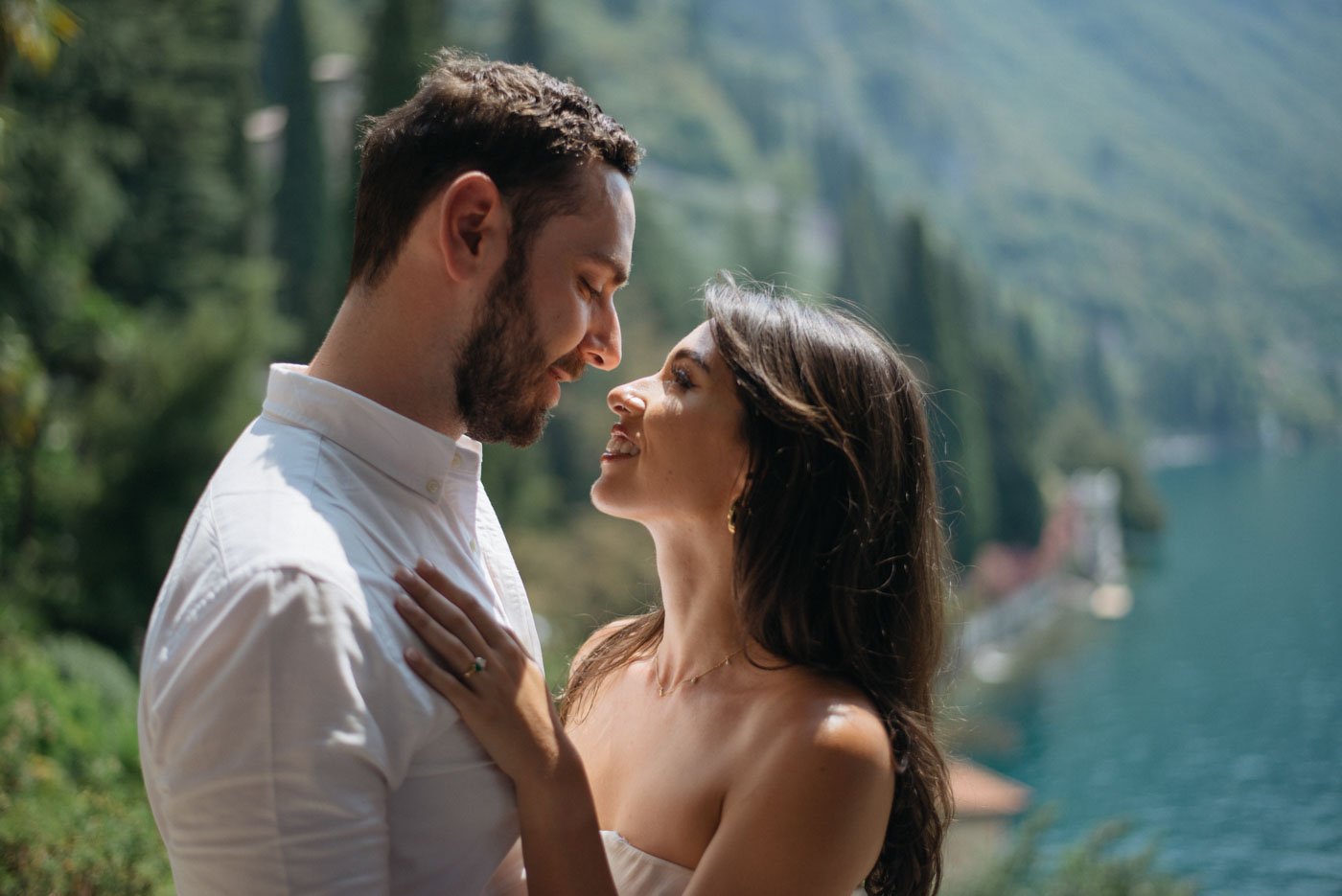 A couple standing close together outdoors near a lake with mountains and trees in the background, gazing into each other's eyes.