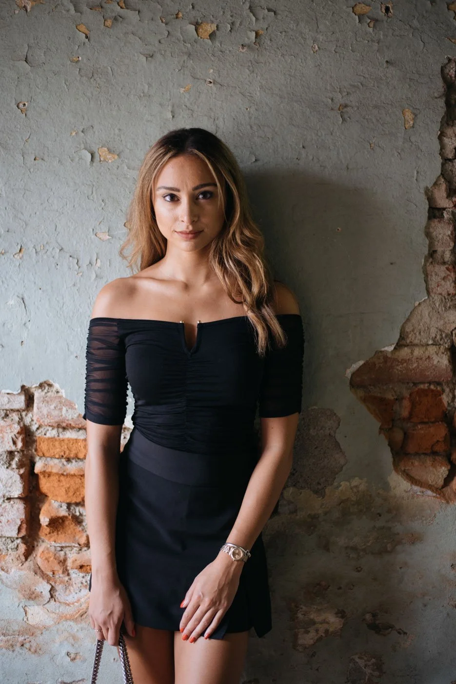 A woman with long, light brown hair wearing a black off-the-shoulder dress standing against a textured wall with exposed brick patches.