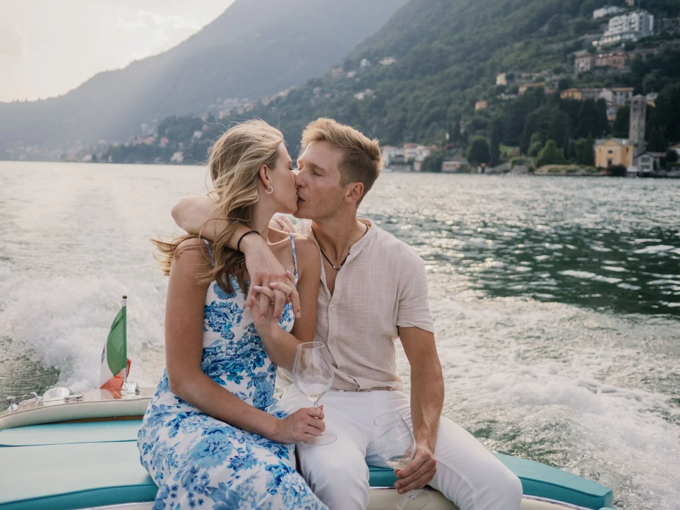 A couple sharing a kiss on a boat with an Italian flag, in a scenic lake setting with mountains and houses in the background.