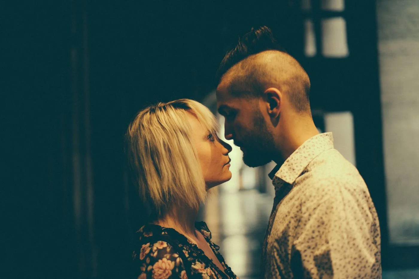 A man and woman face to face in a close, intimate manner indoors with dim lighting.