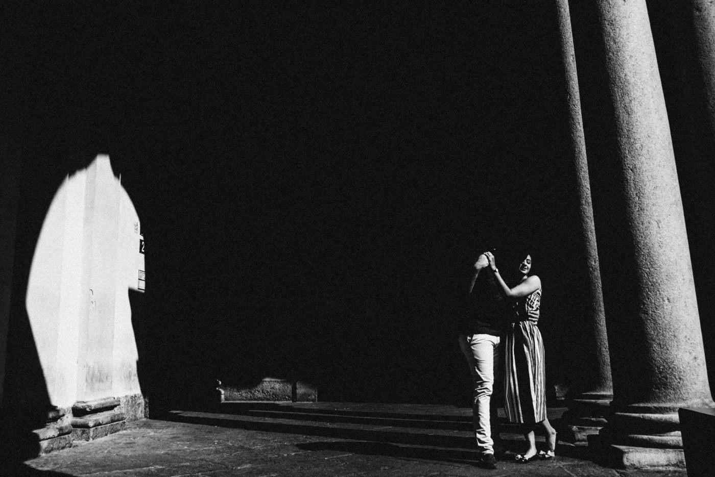 A black and white photo of a couple dancing under a large archway supported by columns, with sunlight casting shadows on the ground.