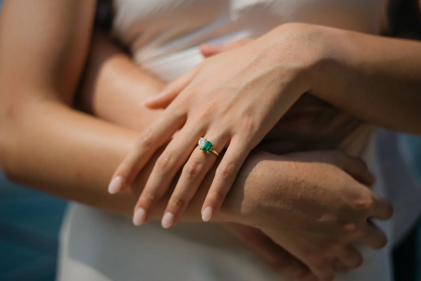 Close-up of a woman's hand wearing a gold ring with an emerald and a diamond, resting on another woman's arm.