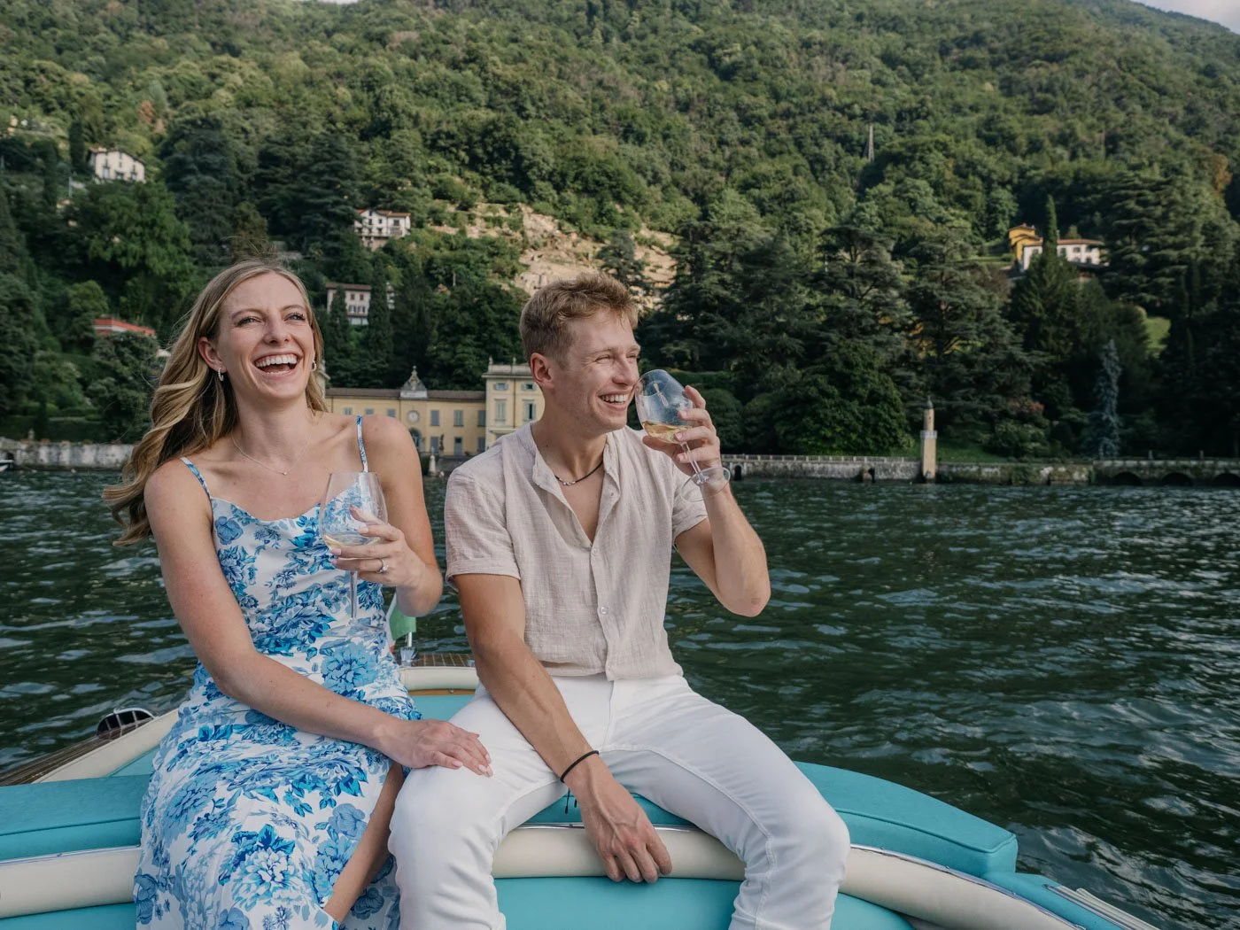 A man and woman enjoying drinks on a boat in front of a scenic lake with lush green hillside and buildings.