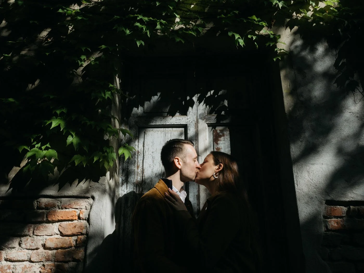 A couple kissing against a rustic wall with green ivy and shadows.