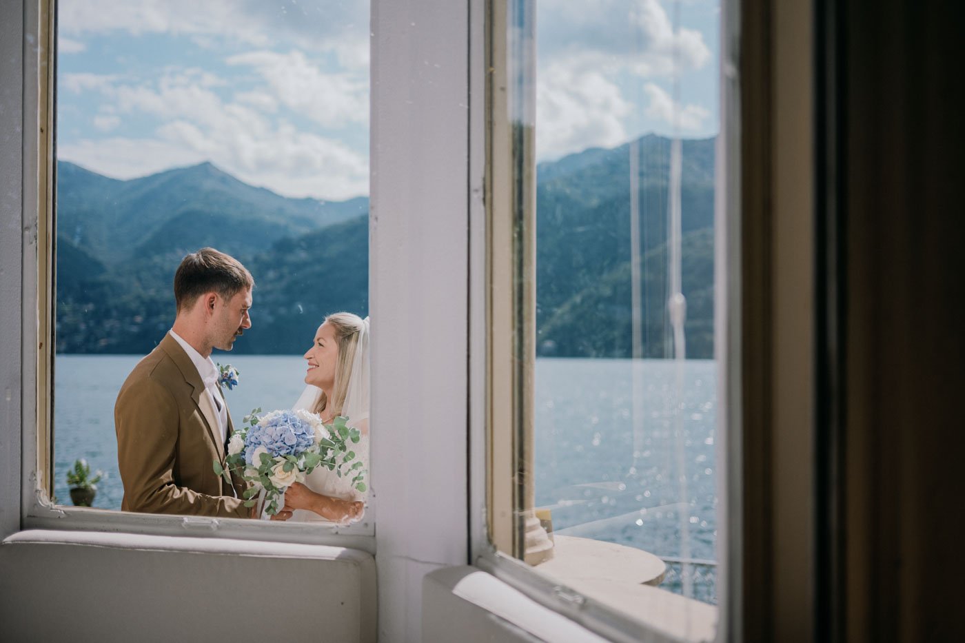 A bride and groom are seen through a window, holding hands and smiling at each other, with mountains and a lake in the background on a sunny day.