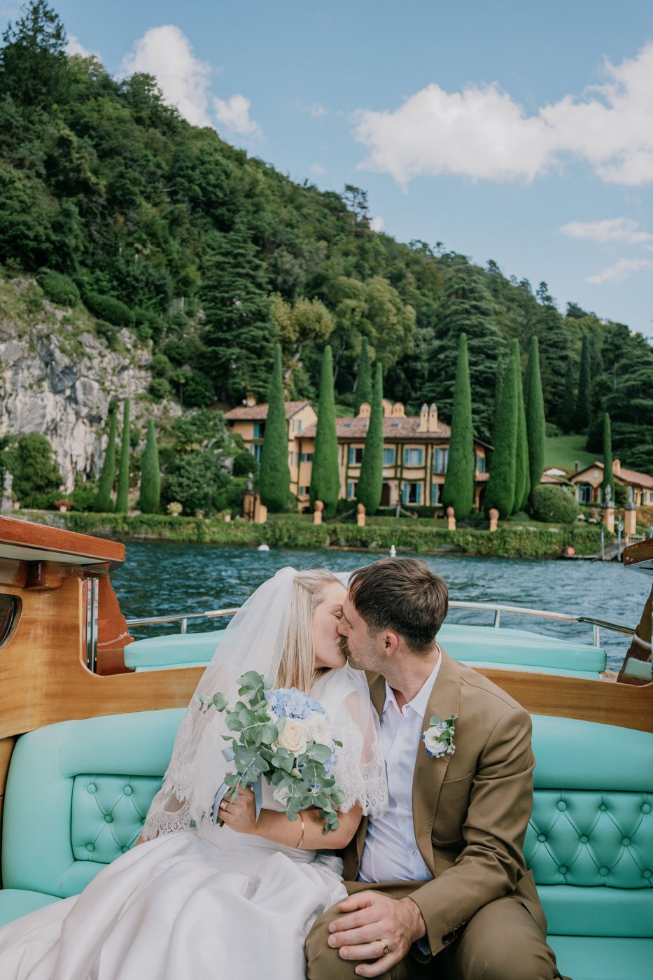 A newlywed couple sharing a kiss on a boat with a scenic backdrop of a lake, green hillside, and tall cypress trees.