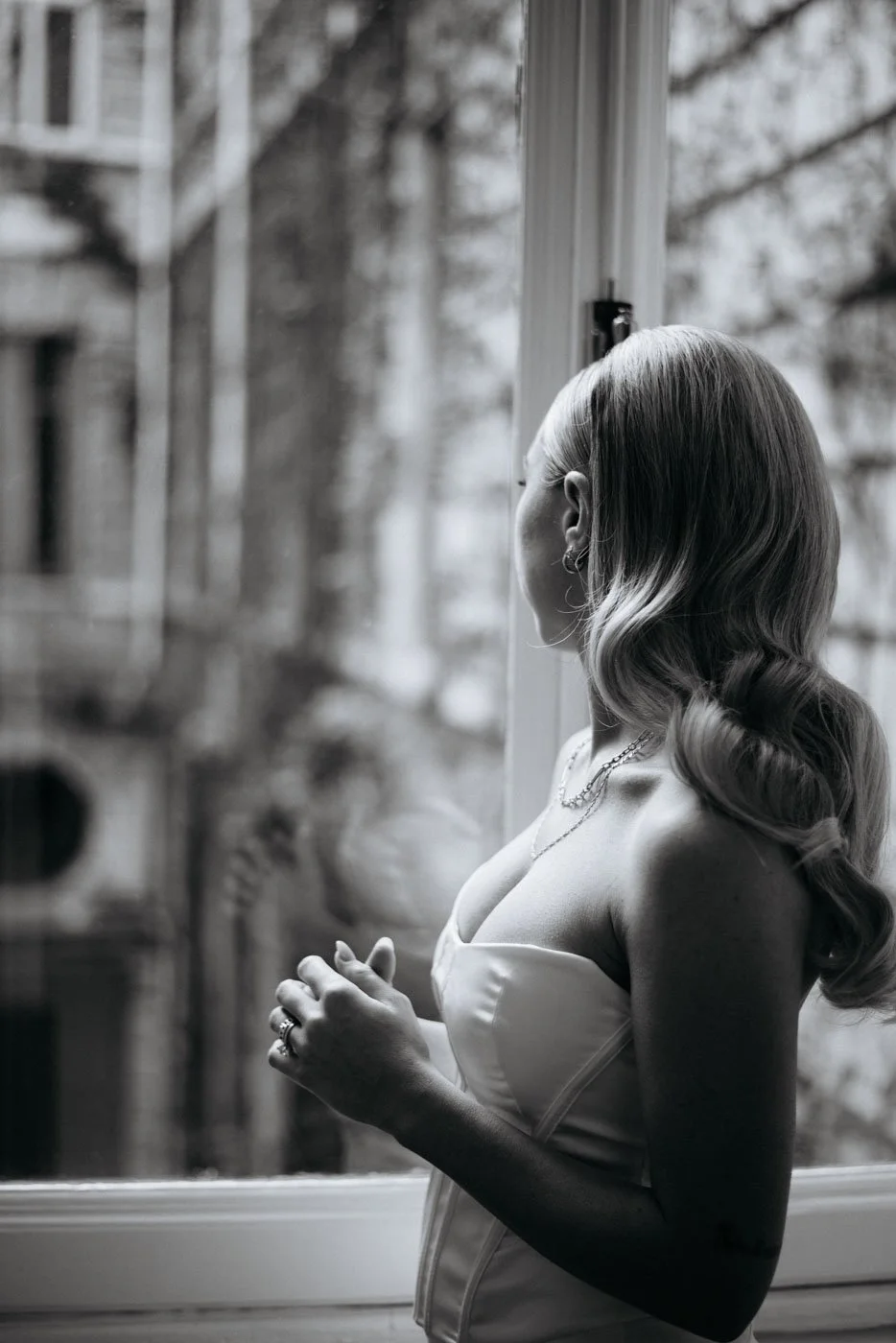 Black and white photo of a woman with long wavy hair looking out a window, dressed in a strapless top, wearing earrings and a necklace.