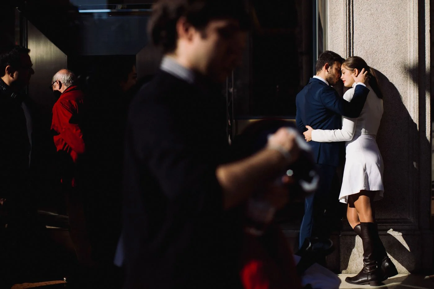 A couple kissing on a city sidewalk, with the woman wearing a white dress and knee-high boots, and the man in a dark suit. Other people are walking by, some wearing masks, and a man in the foreground is looking at his phone.