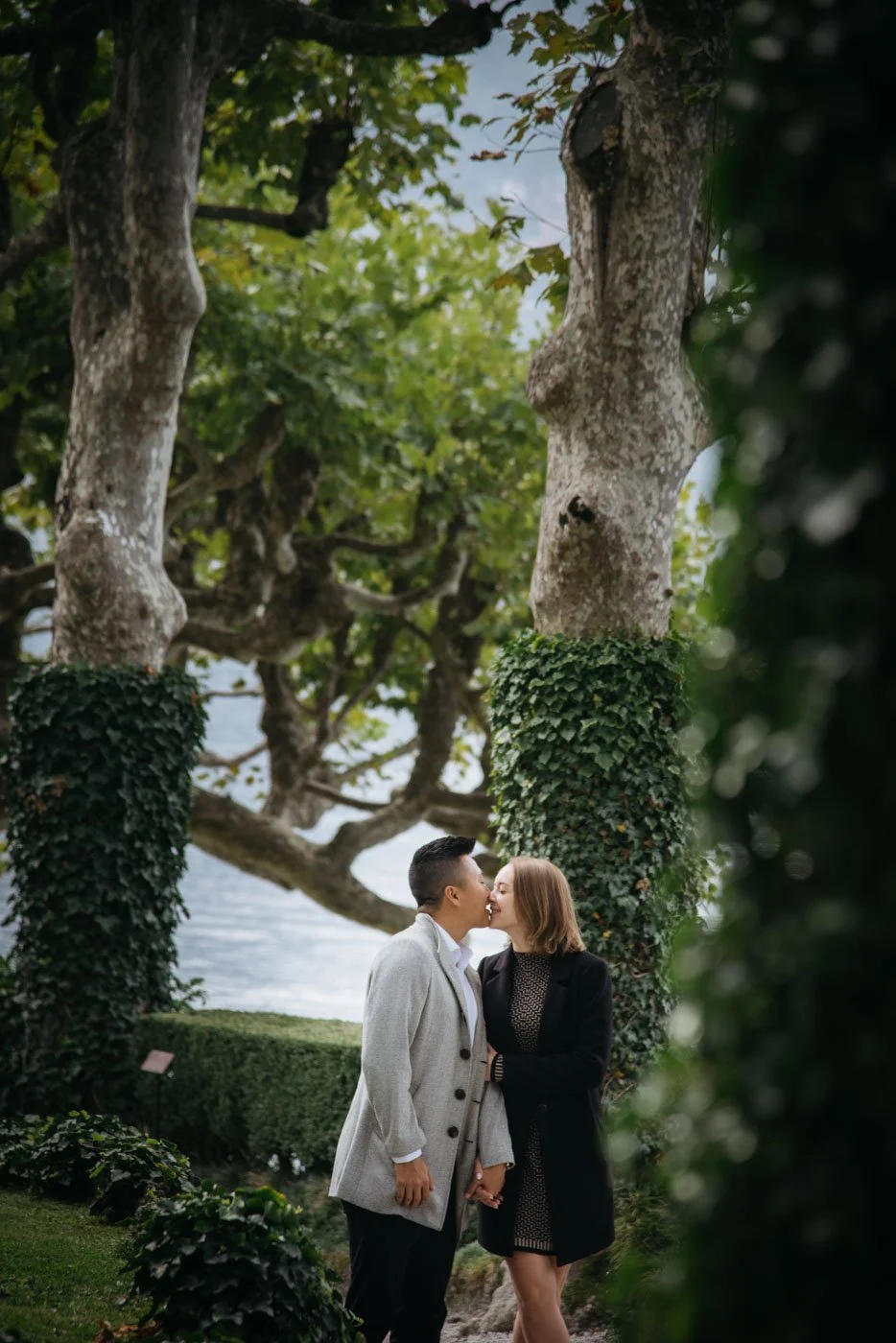 A couple kissing outdoors amid lush greenery, trees, and ivy-covered trunks near a body of water.