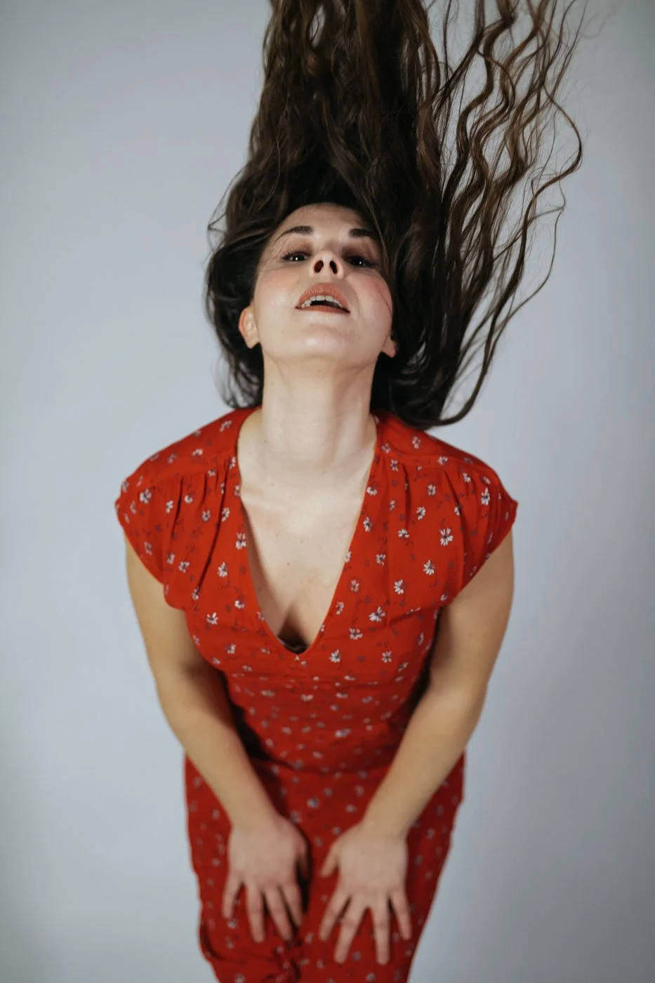 A woman with long, curly brown hair is leaning forward, looking at the camera, with her hair flowing upward. She is wearing a red dress with small white flower patterns. The background is plain and light-colored.