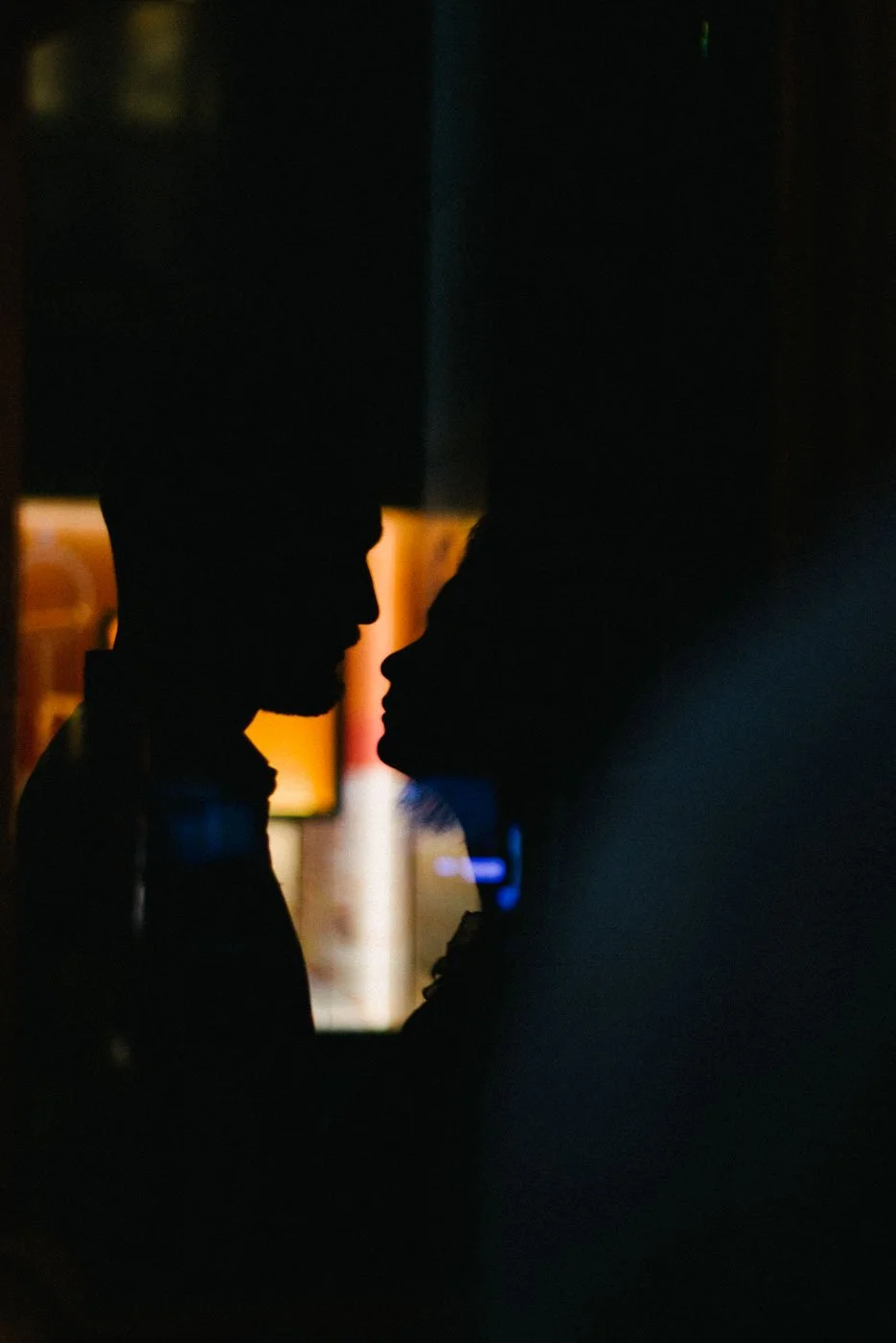 Silhouette of a man and woman facing each other closely, with a blurred background of colorful lights, in a dark setting.